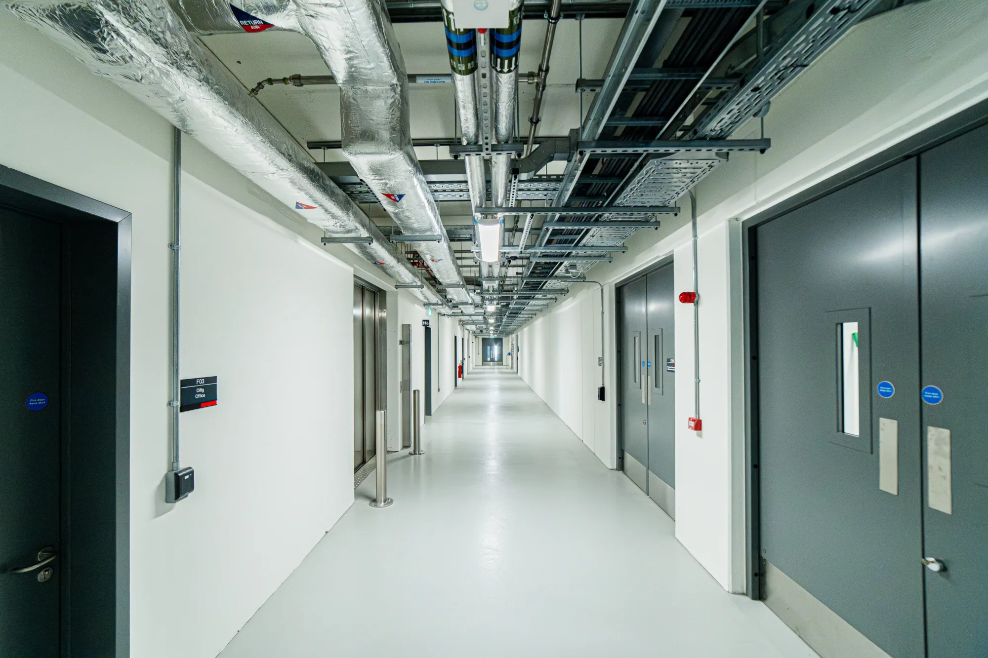 A long, well-lit corridor with white walls, light grey floor, and exposed ceiling pipes and cables. Grey doors with small windows line both sides.