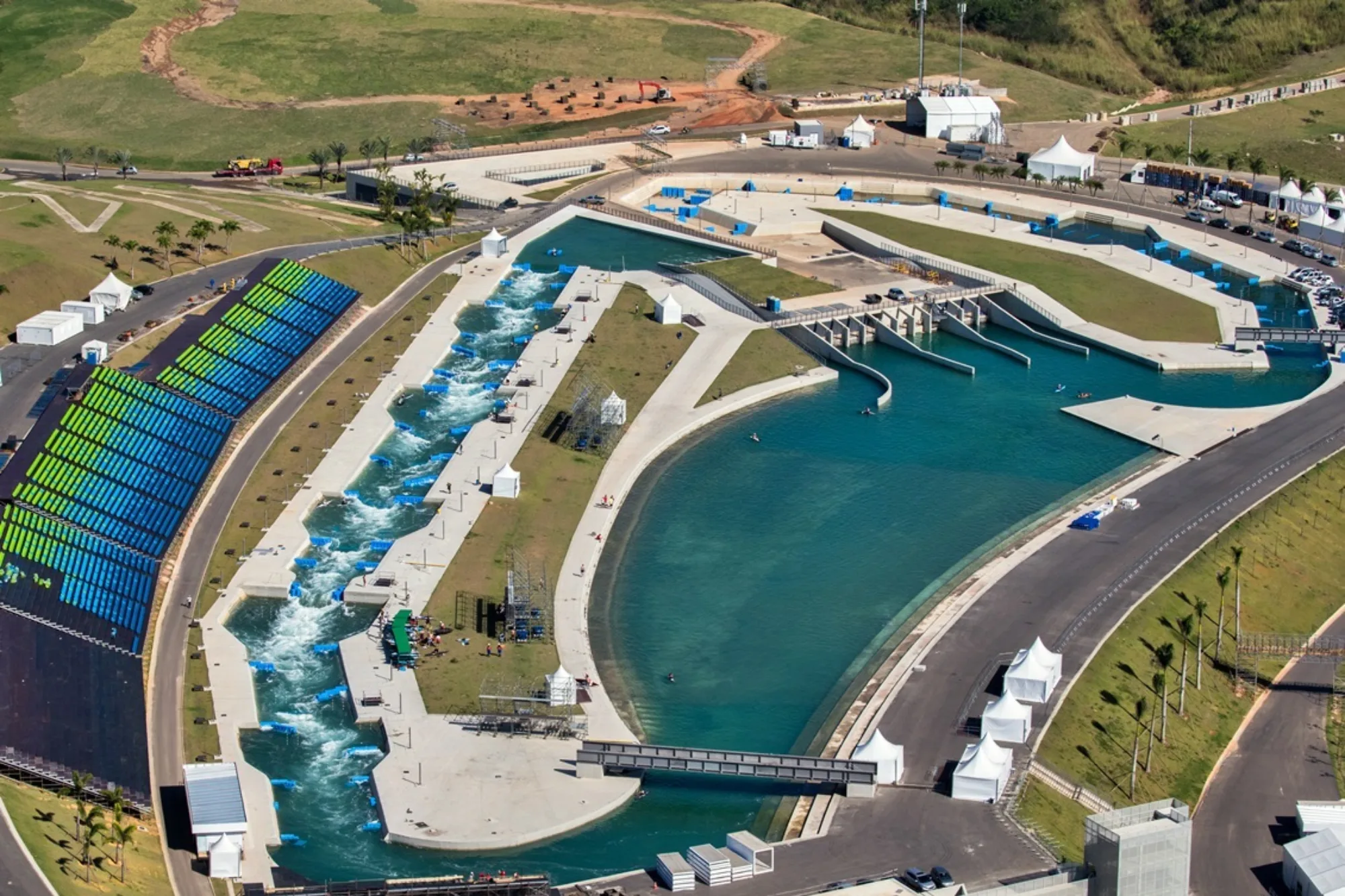 Aerial view of a large white‑water sports venue with rapids, a calm water basin, walkways, bridges, seating stands, tents, and surrounding grassy areas and roads.