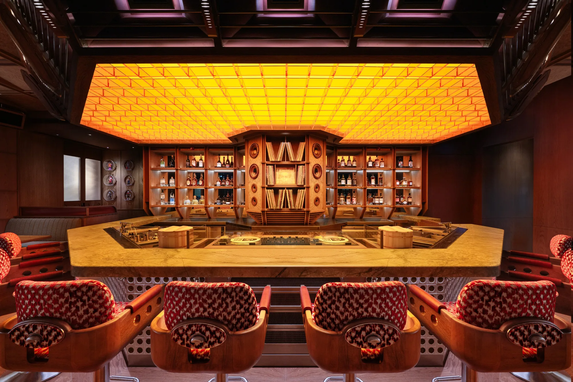 Interior of a bar with patterned upholstered chairs facing a large stone counter, illuminated by a glowing yellow ceiling panel, with shelves behind the bar displaying bottles and decorative objects.