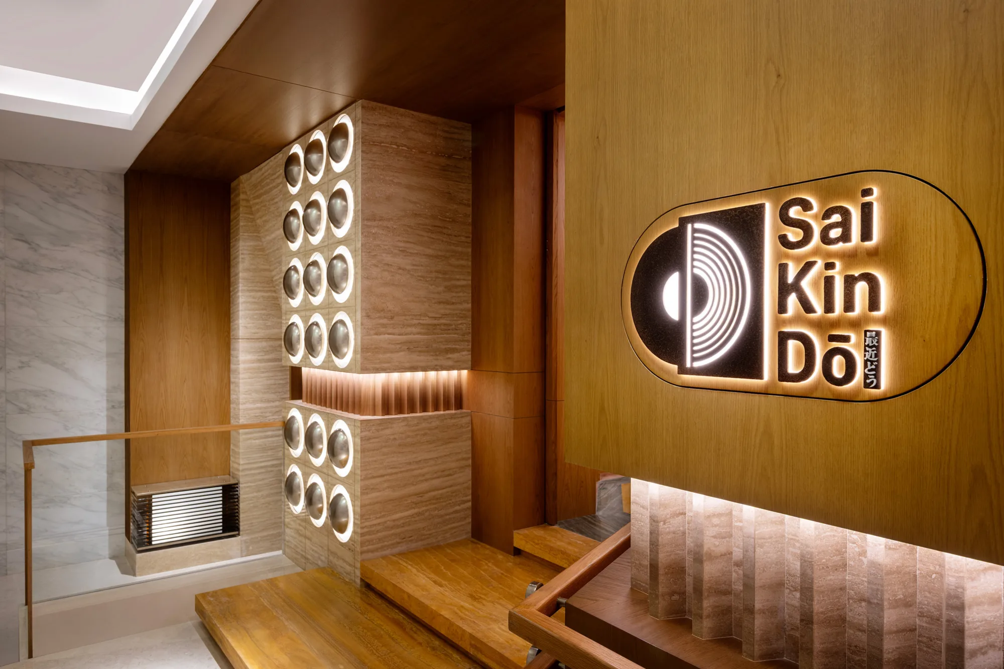 Interior hallway with wood and marble surfaces, illuminated circular wall lights, and a wooden sign displaying the text ‘Sai Kin Dō’ beside a graphic symbol, positioned above a staircase.