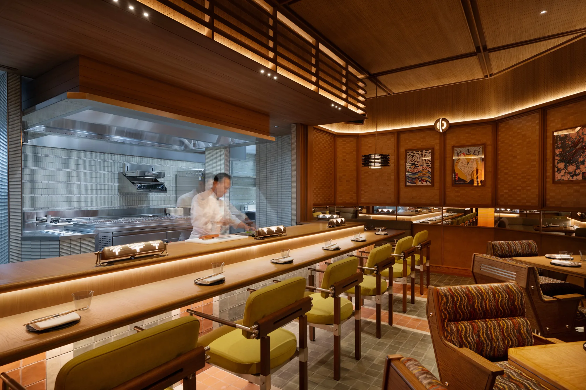 Interior of a restaurant with a long wooden counter lined with green‑cushioned chairs, a chef preparing food behind a glass partition, and framed artwork on wood‑panelled walls under warm lighting.