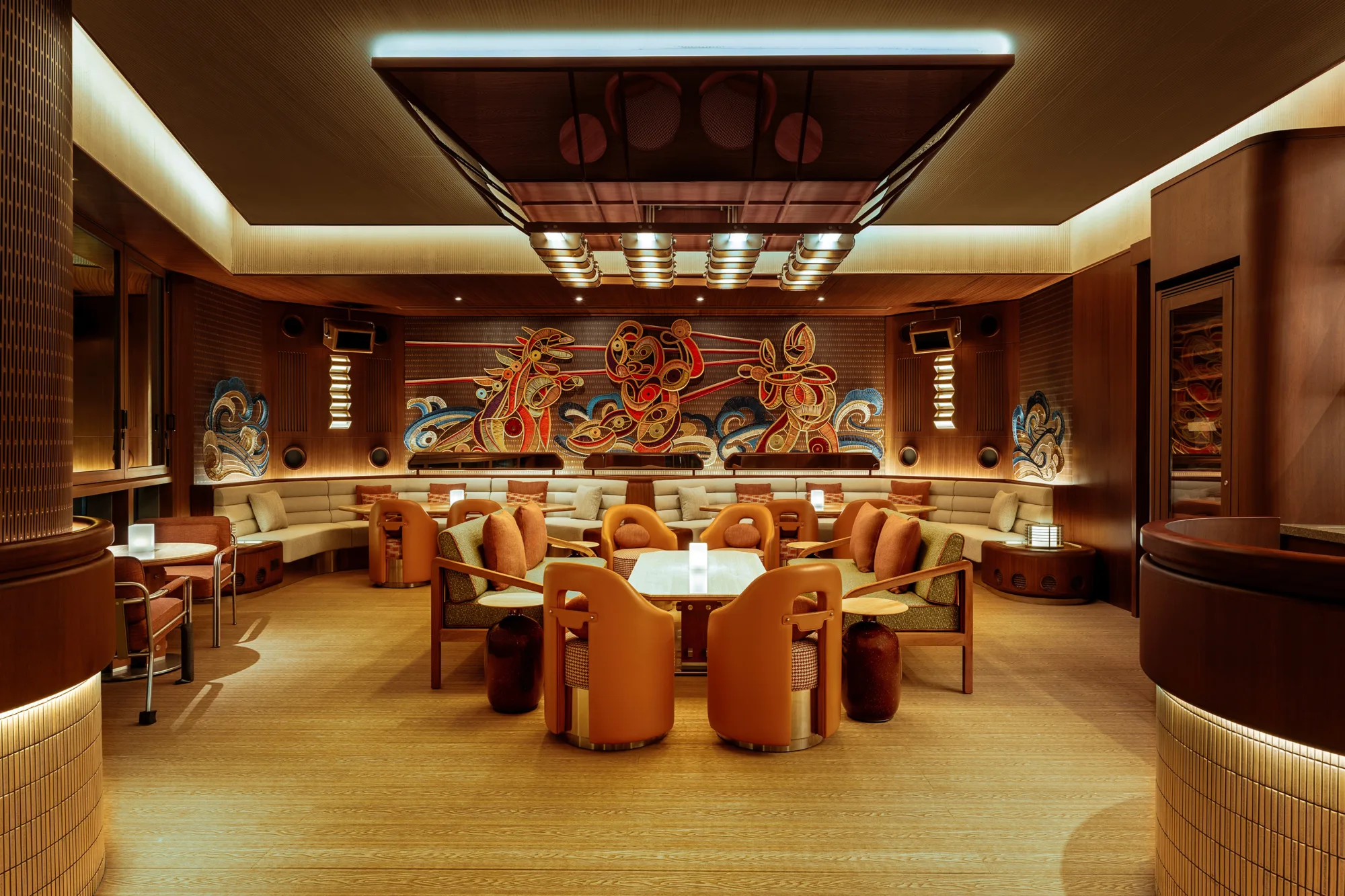 Interior of a lounge with wooden flooring, curved seating, and orange chairs around tables, featuring a large colourful mural of stylized figures on the back wall and warm recessed lighting throughout.