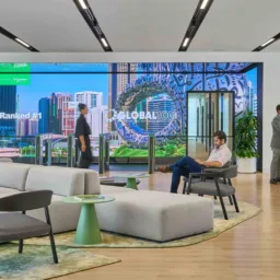 A modern office lobby with contemporary seating, plants, and a large digital wall display showing city imagery and the text ‘Ranked #1’ and ‘Global 100’. Several people sit, walk, or speak at a reception desk.