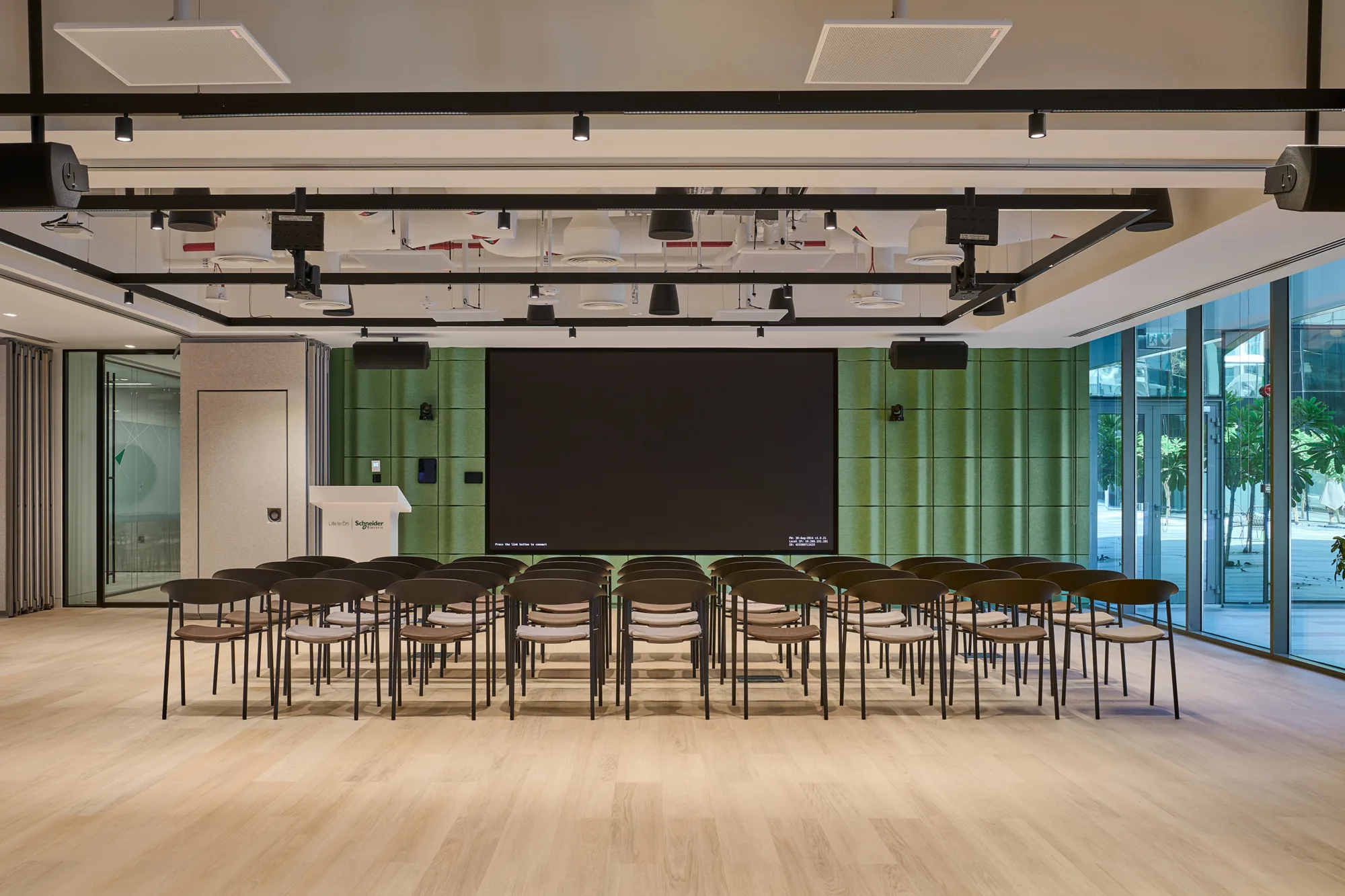 Modern conference room with green panel walls, wooden flooring, and large windows on the right. Rows of empty chairs face a large black screen at the front, with a white podium to the left and ceiling-mounted equipment above.