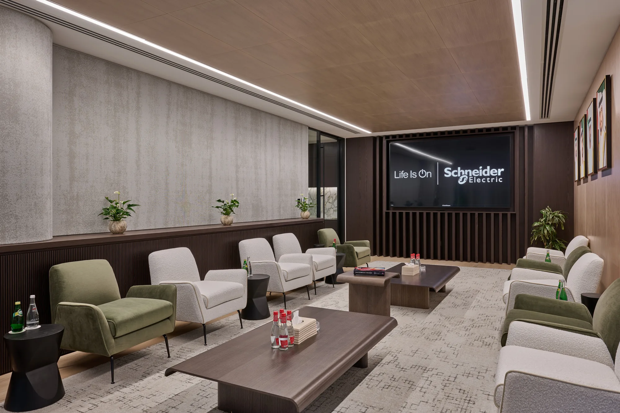 Modern waiting area with wooden ceiling, grey walls, green and grey chairs, and two wooden coffee tables holding magazines, bottled water, and small plants. A large screen displays "Life Is On" and "Schneider Electric."
