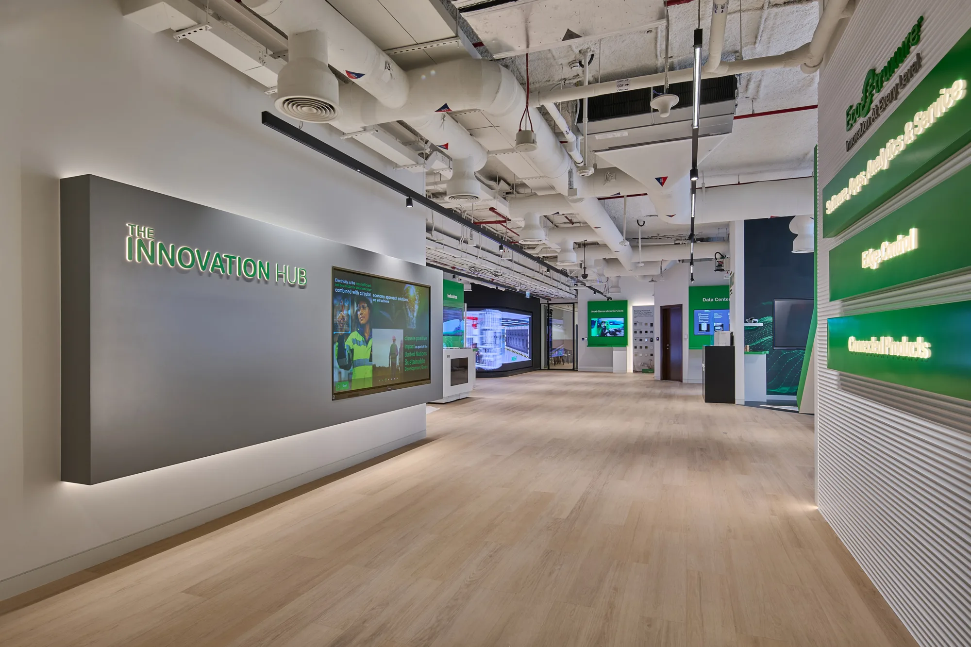 Spacious room with light wood flooring and exposed ceiling pipes. Left wall has a sign reading "The Innovation Hub" and a screen showing a person in safety gear. Right wall displays green signs with text including "Edge Control" and "Connected Products."