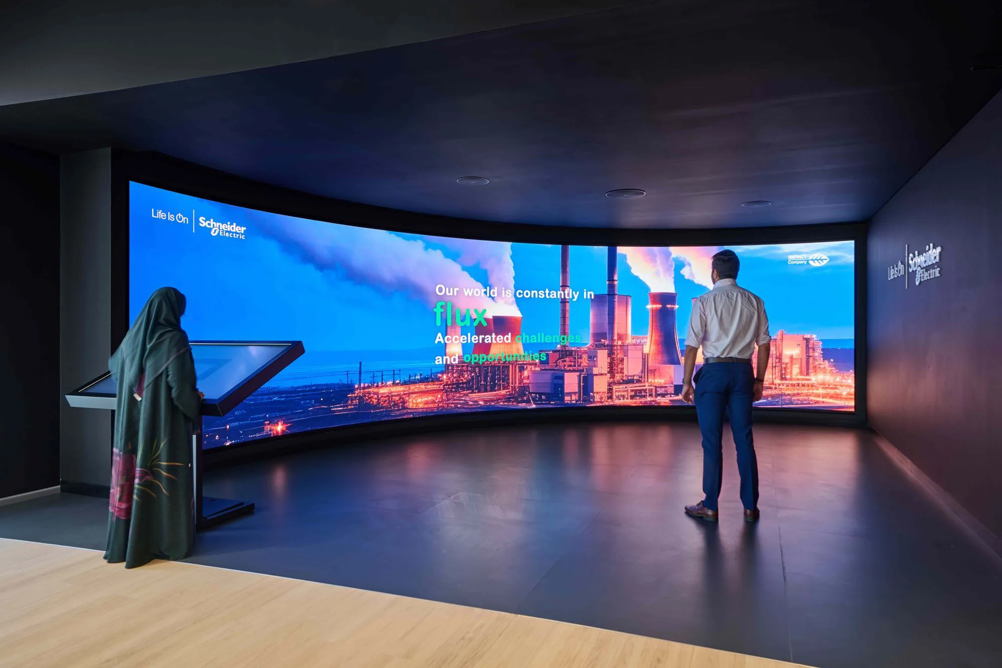 A curved digital wall displays an image of an industrial facility with text about a world in flux. One person stands viewing the screen while another uses an interactive touchscreen in a dark exhibition-style room.