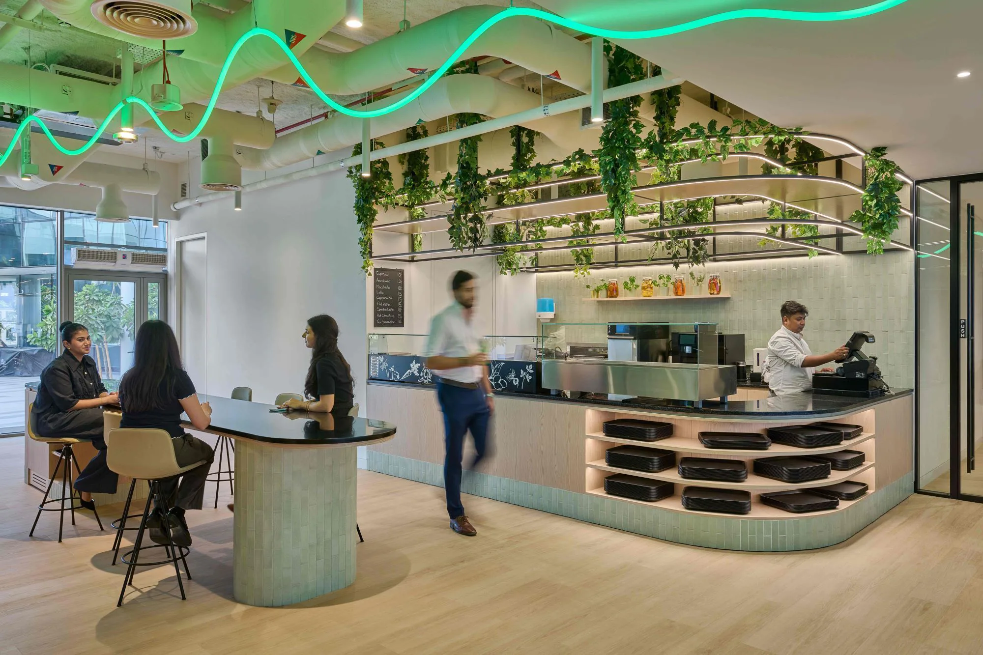 A modern café-style area with a counter, coffee machines, and hanging greenery. Three people sit at a high table while another walks past. A staff member works behind the counter beneath curving neon lights.