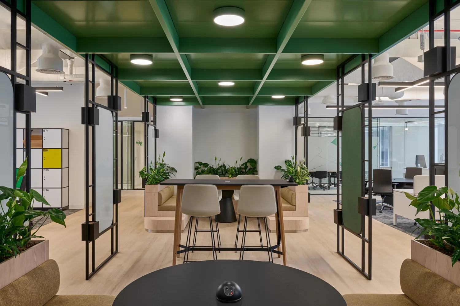Modern office space with green ceiling, wooden floors, and glass partitions. Foreground has a round black table with a small device. Centre features beige chairs at a high table, surrounded by plants. Lockers and open office area in background.