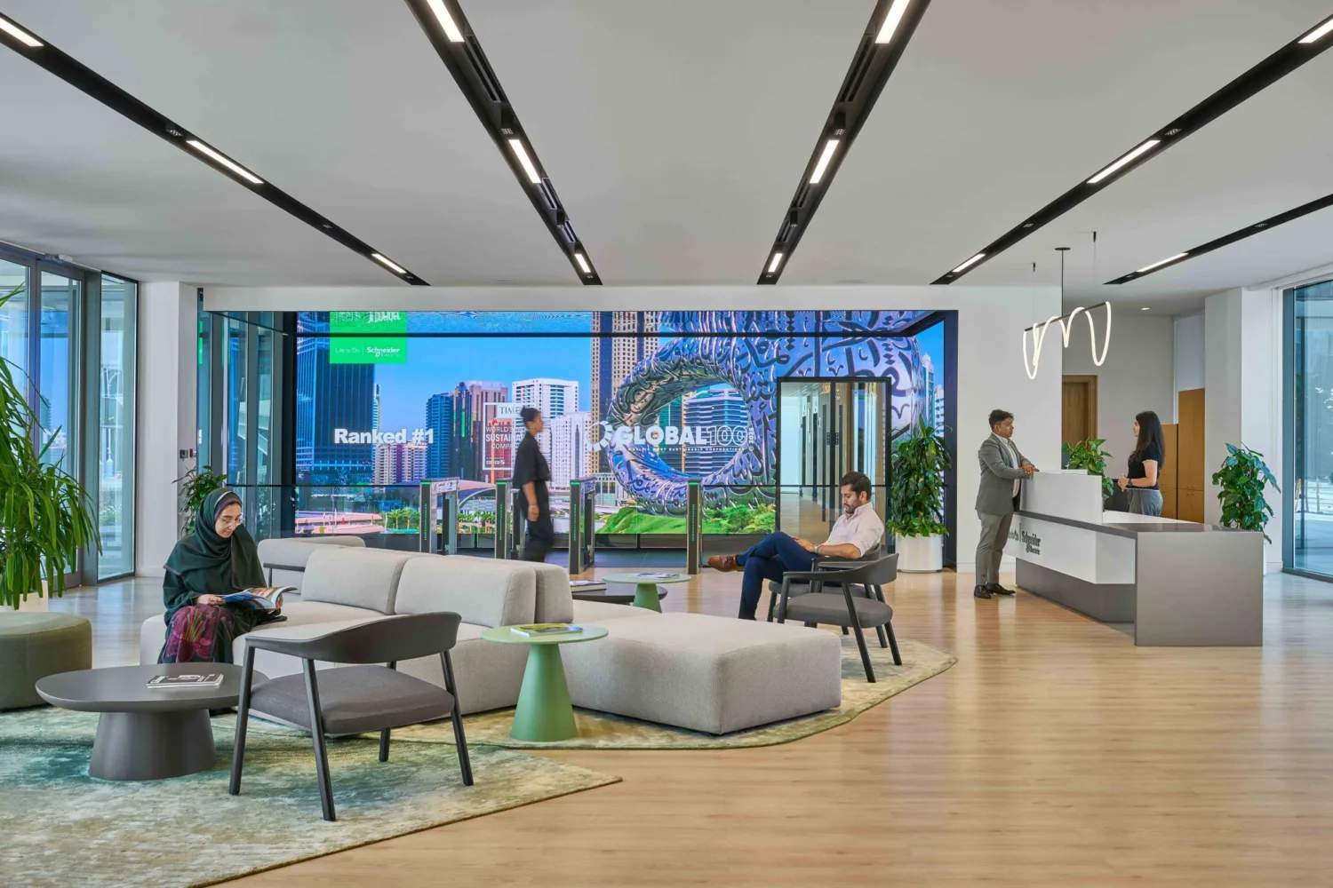 A modern lobby with lounge seating, low tables, and large digital screens showing city visuals. Several people sit, walk, or speak at a reception desk. Floor‑to‑ceiling windows and plants brighten the space.
