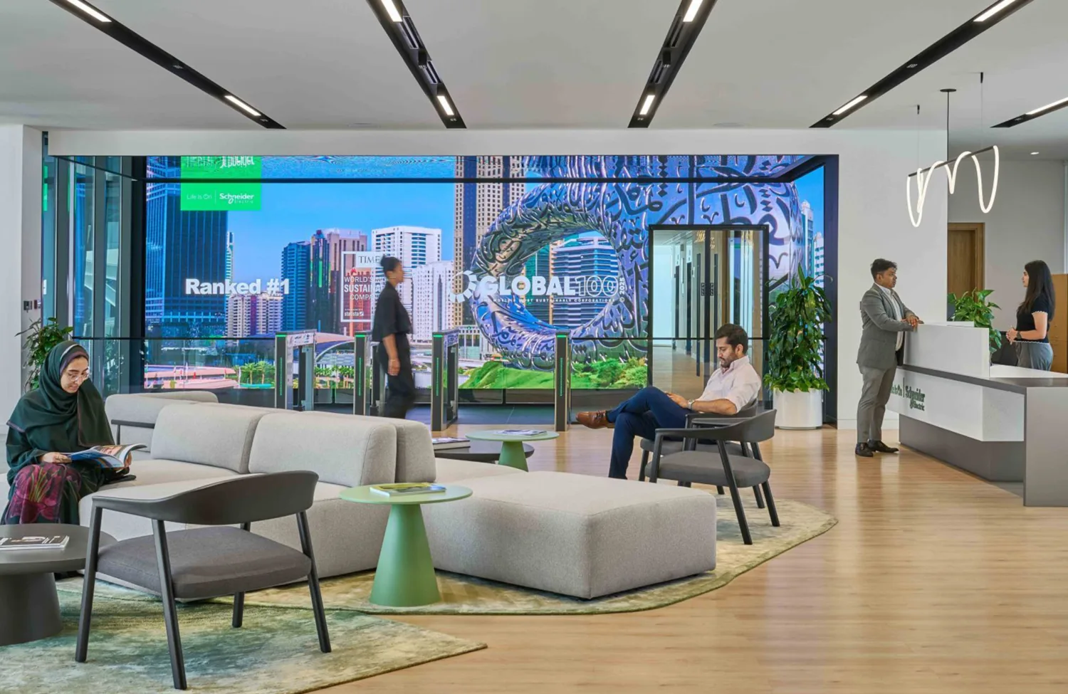 A modern office lobby with contemporary seating, plants, and a large digital wall display showing city imagery and the text ‘Ranked #1’ and ‘Global 100’. Several people sit, walk, or speak at a reception desk.