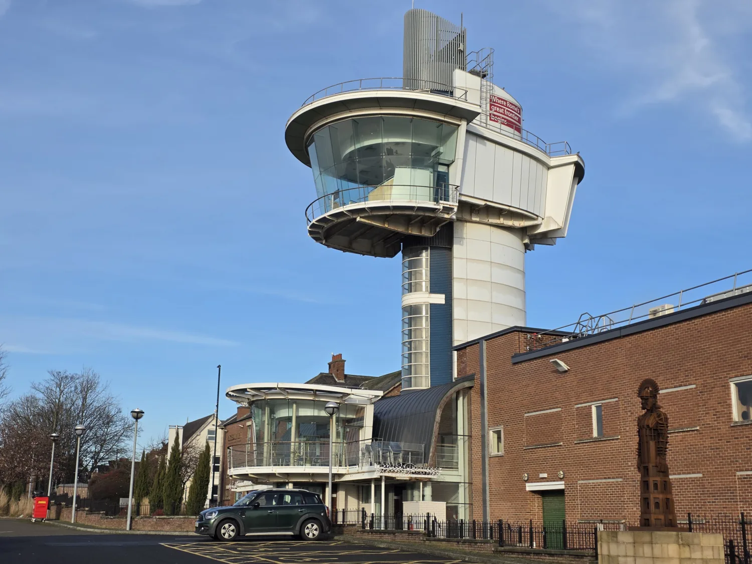 A tall observation‑tower structure with a circular glass viewing area rises behind a modern brick building. A parked car, a metal statue, streetlamps, trees, and a blue sky are visible in the scene.