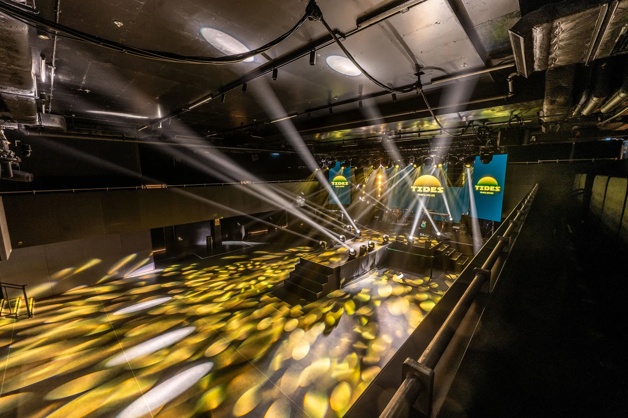 Large indoor event venue with a stage illuminated by multiple bright spotlights and yellow patterned floor lighting. Two vertical screens on either side of the stage display the word “TIDES” in bold yellow letters on a blue background. Overhead rigging and lighting equipment are visible.