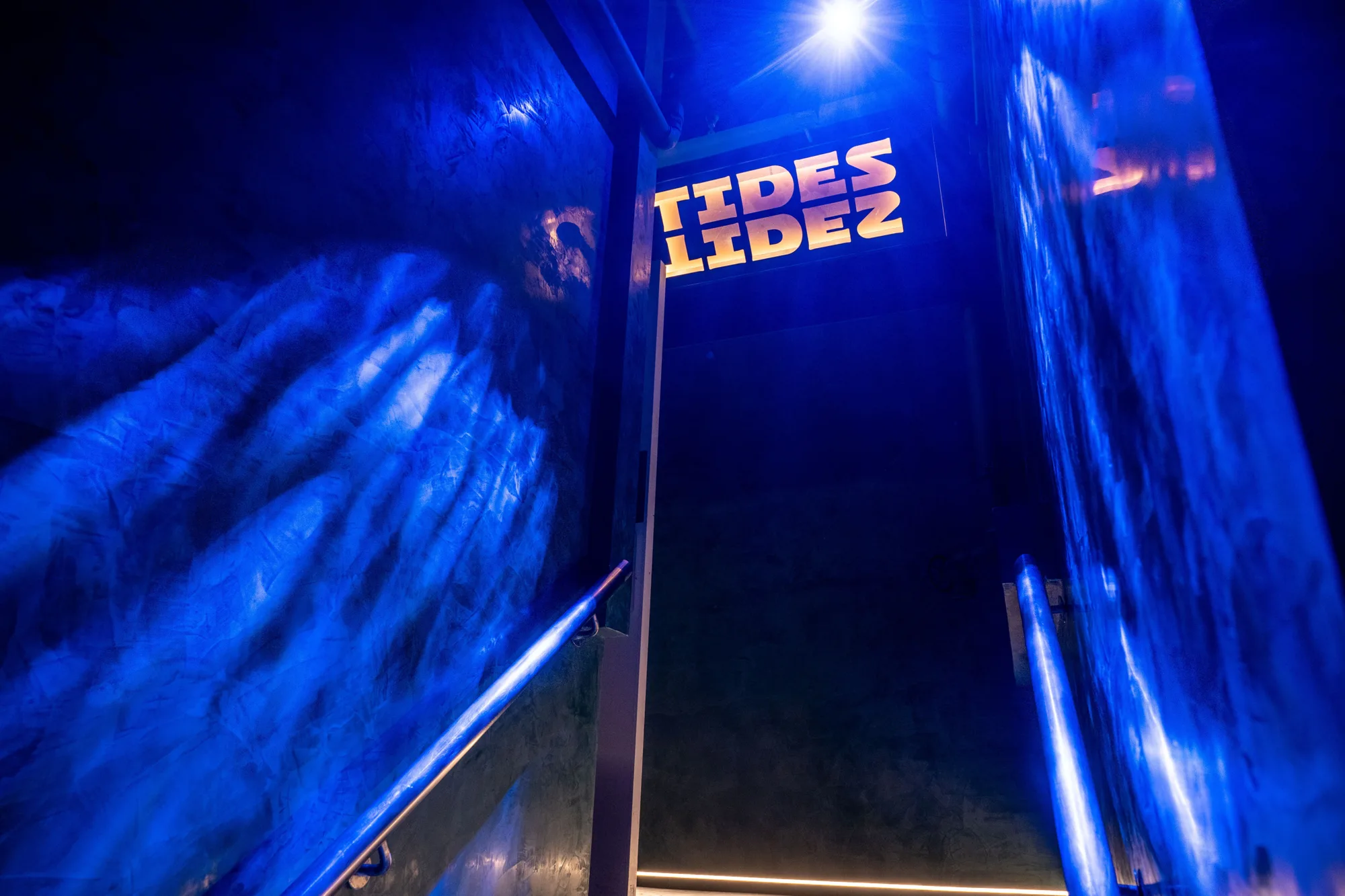Dimly lit stairway with textured blue walls and metal handrails leading upward to a sign displaying the word “TIDES” in bold yellow letters on a dark background. A bright light source is visible above the sign.