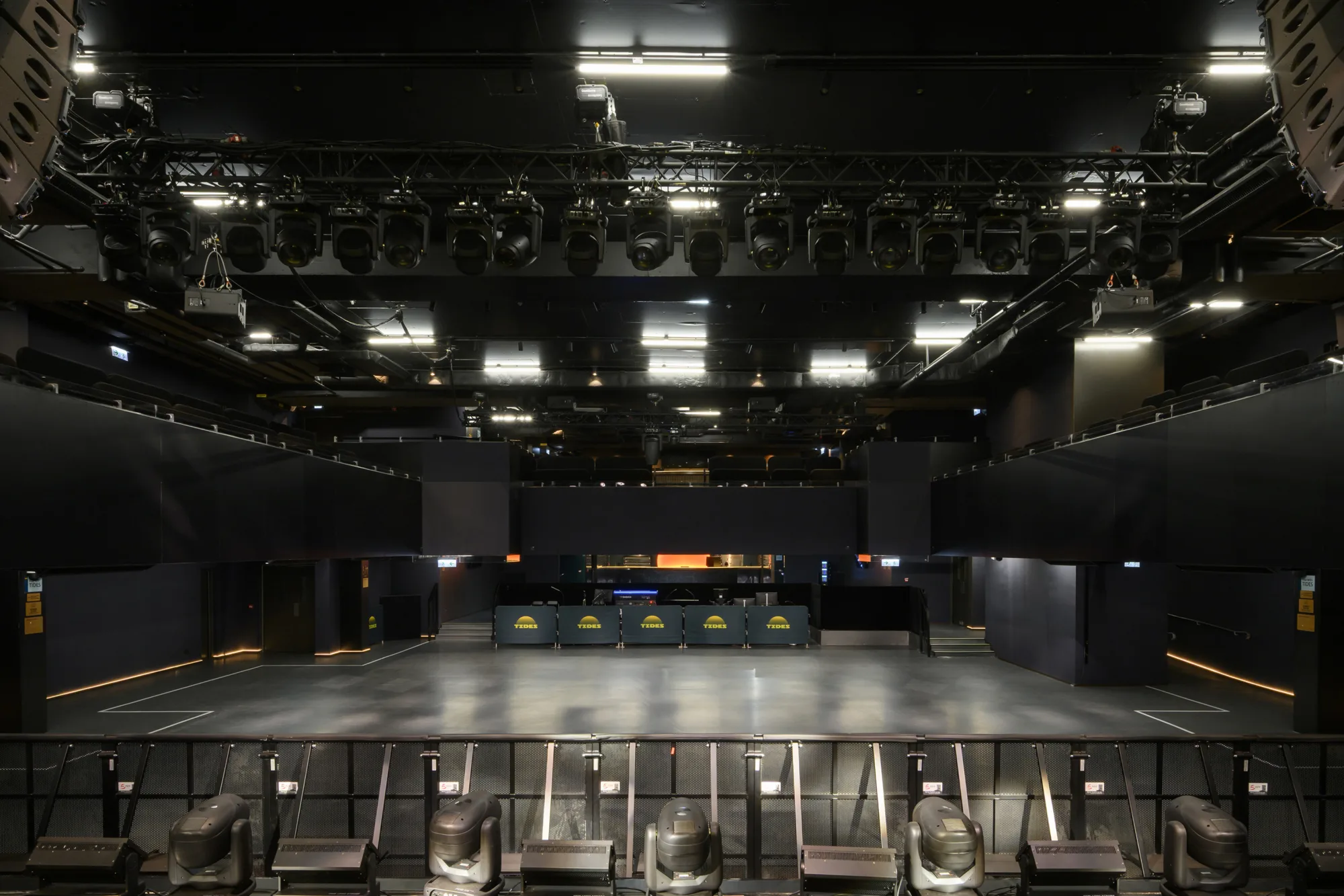 A large indoor venue with an empty open floor faces a bar area at the back displaying three screens showing a yellow sun‑shaped graphic and the word “TIDES.” Overhead rigging, lighting equipment, and tiered seating surround the space.