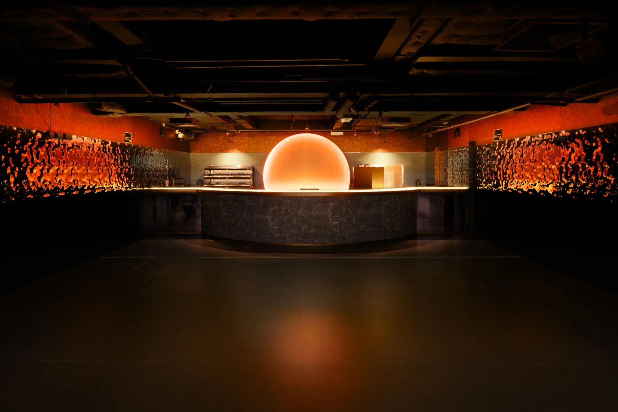 A bar counter with a stone-textured front sits beneath a dark ceiling. A large circular orange light feature glows behind the counter, illuminating surrounding textured wall panels with warm amber lighting.