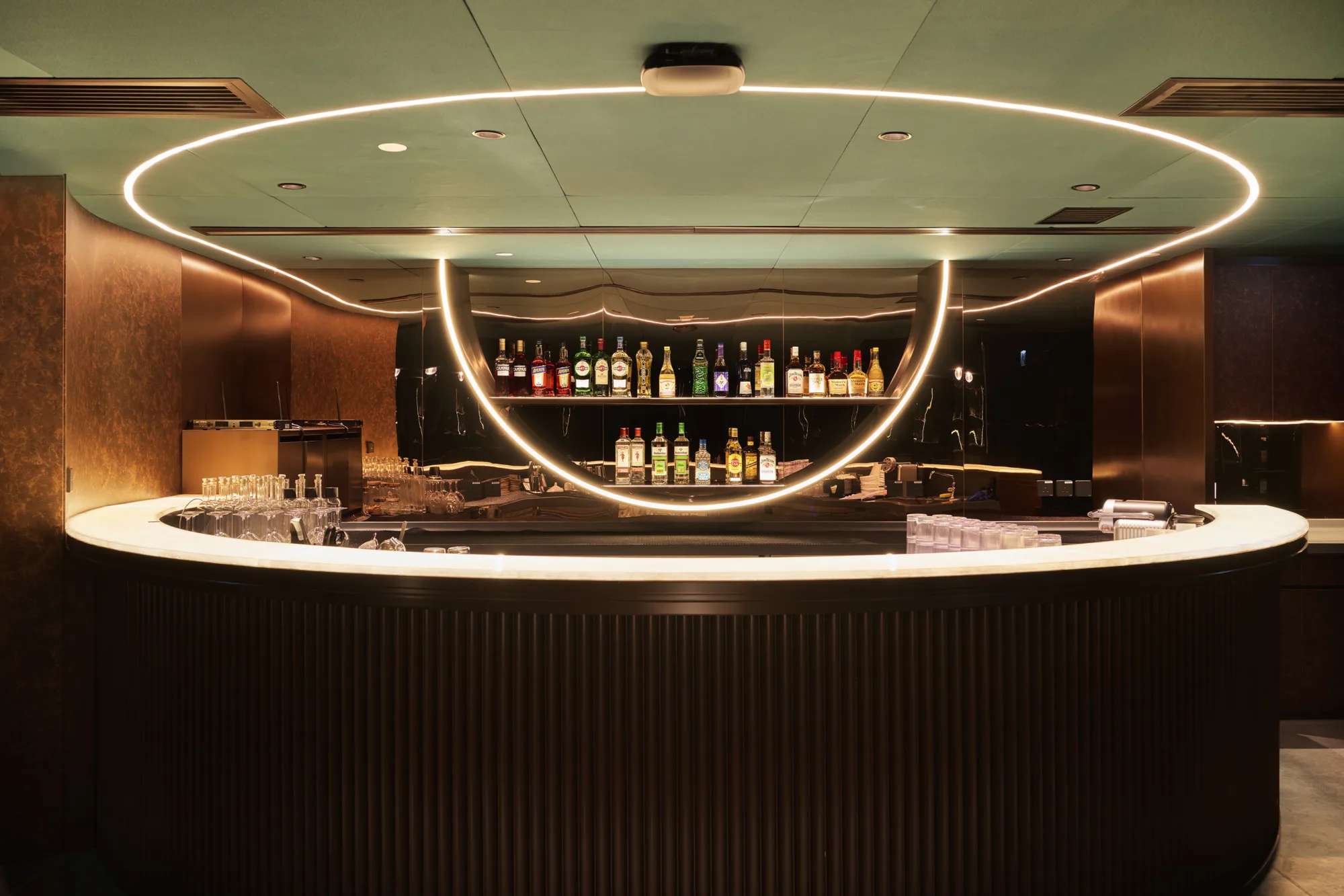 A curved bar with a light‑coloured countertop stands beneath a ceiling with two large circular light rings. Shelves behind the bar display bottles of various colours, with glasses and equipment arranged on the counter.