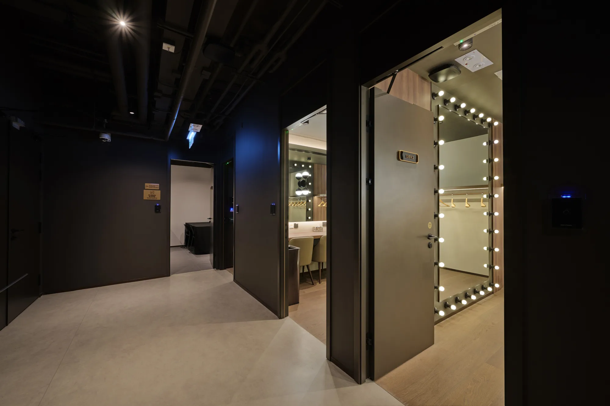 A dim corridor with dark walls leads to two open rooms. One room contains a counter with chairs and illuminated mirrors framed by small round bulbs. The other room has an open doorway with hanging rails and lights.