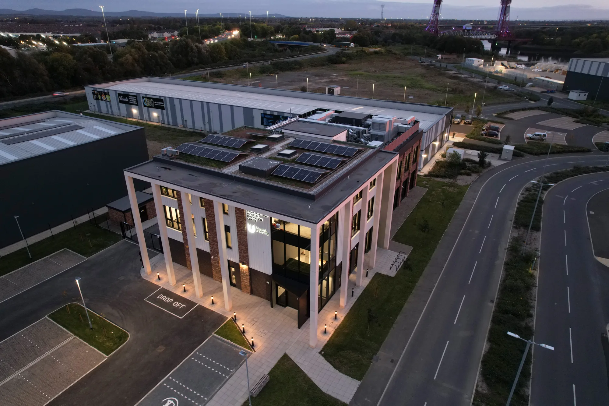 Aerial view of a modern multi-storey office building with rooftop solar panels, surrounded by parking lots and roads. Exterior lights illuminate the building. Industrial structures and greenery are in the background.