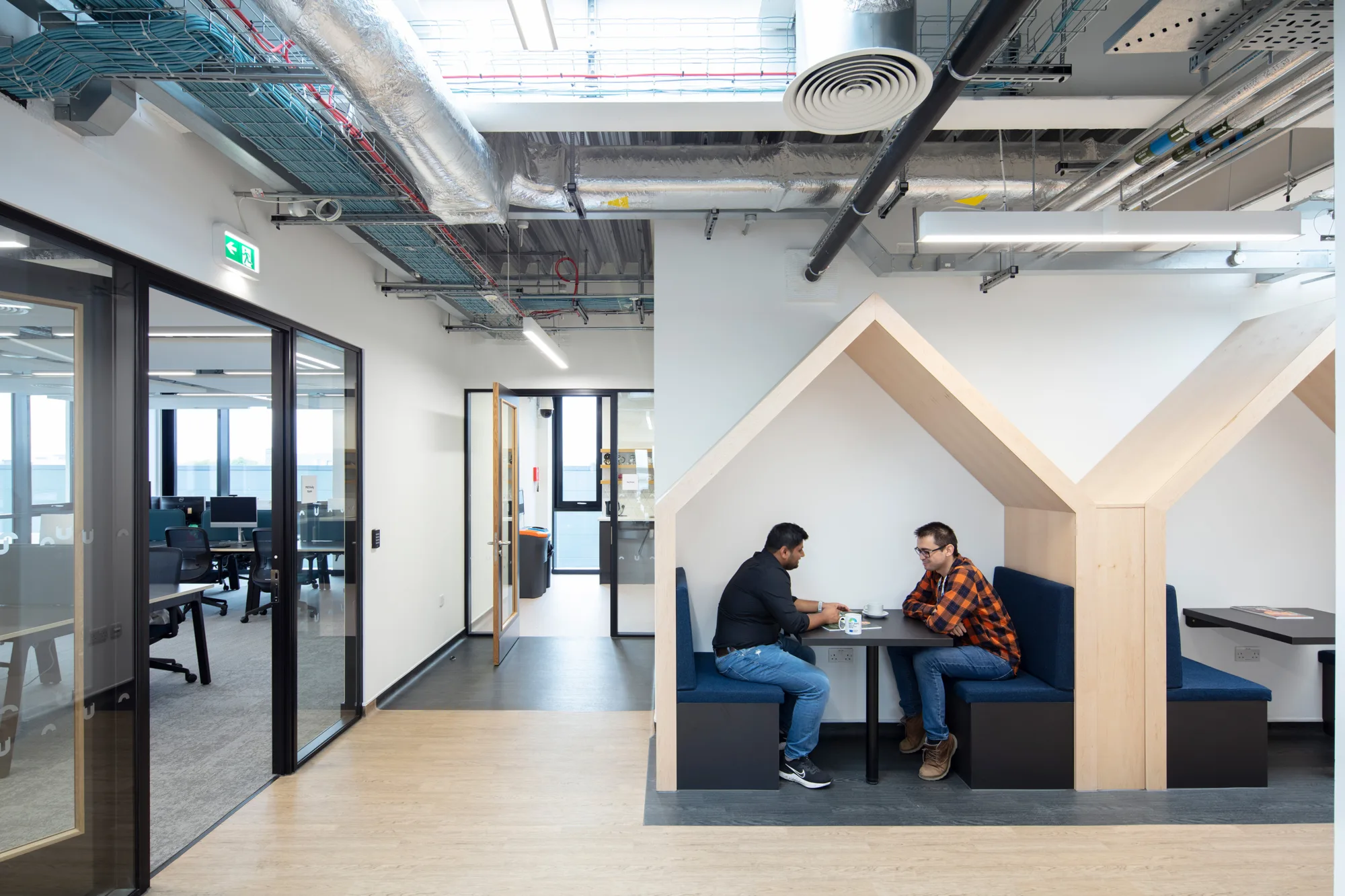 Two people sit at a table inside a wooden, house-shaped booth with blue seating in a modern office. The space features exposed ceiling ductwork, glass-walled meeting rooms, and an emergency exit sign.