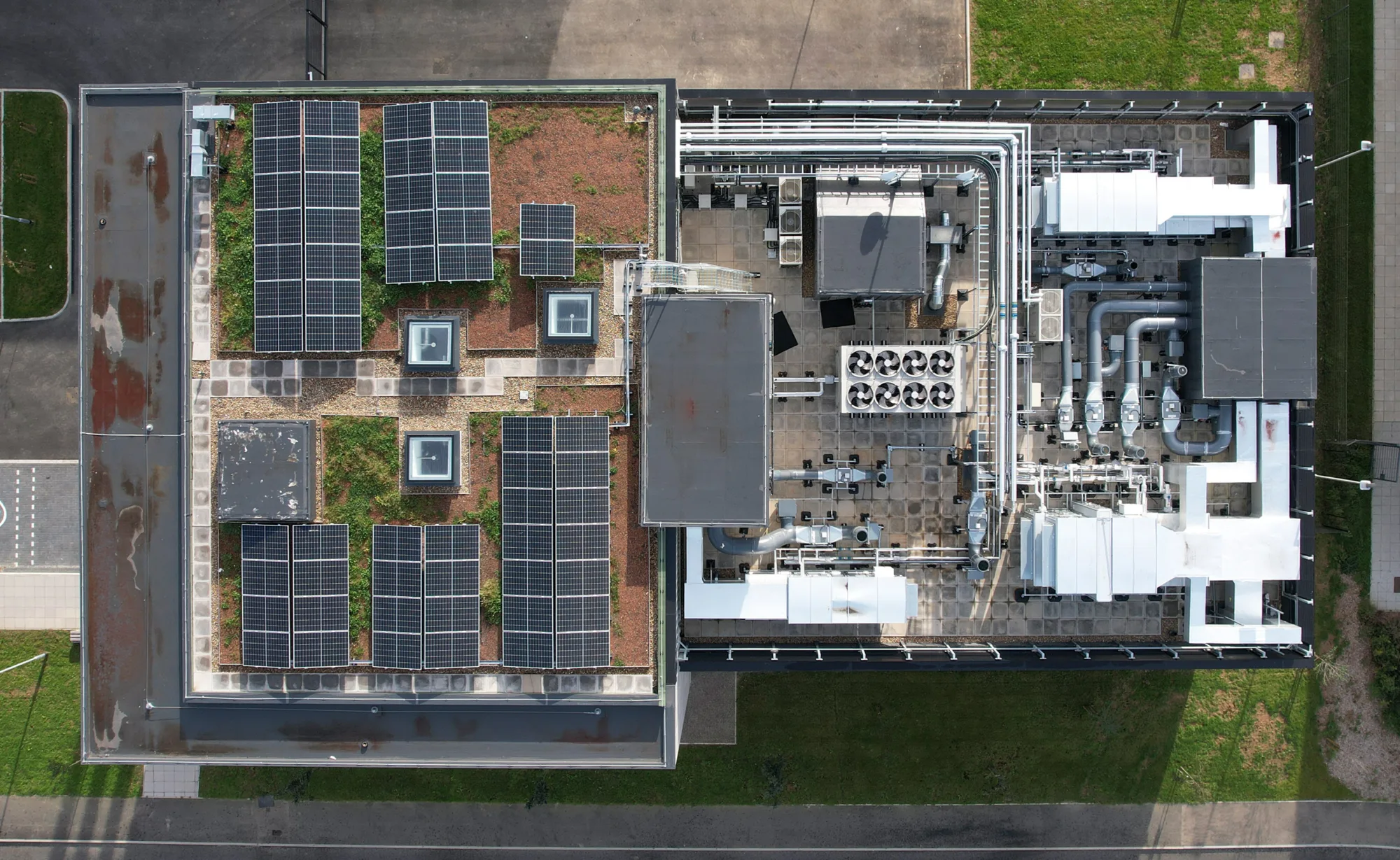 Aerial view of a rooftop with solar panels and green patches on the left, and industrial equipment including pipes and ventilation units on the right.