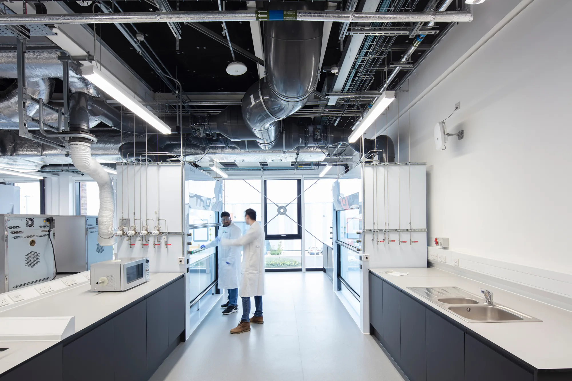 Two people in lab coats stand in a modern laboratory with white walls, black cabinets, stainless steel countertops, a microwave, sinks, gas lines, and large windows providing natural light.