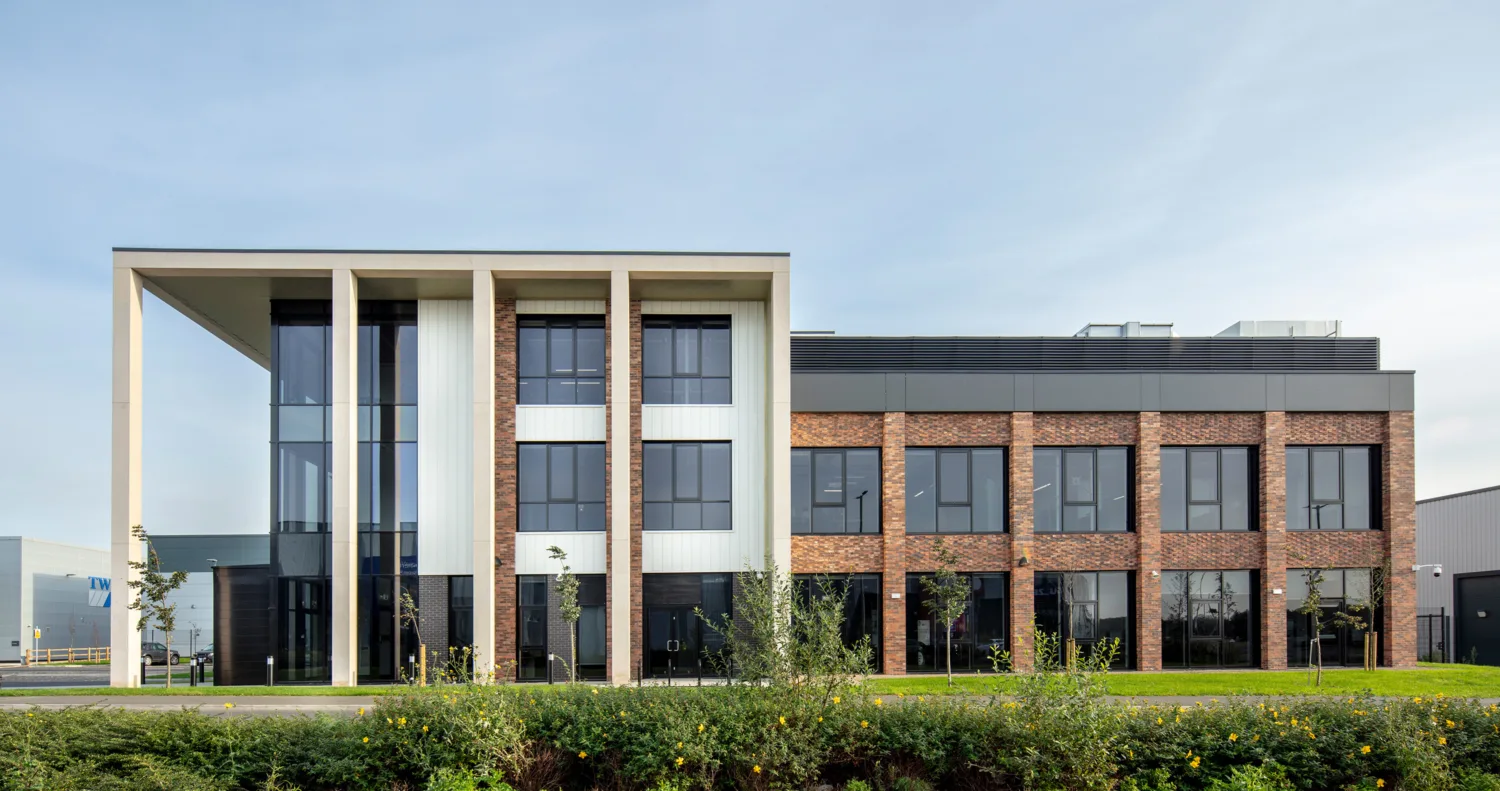 Modern three-story building with brick and white panel exterior, large windows, and an overhanging structure supported by columns. Surrounded by greenery with a clear sky in the background.