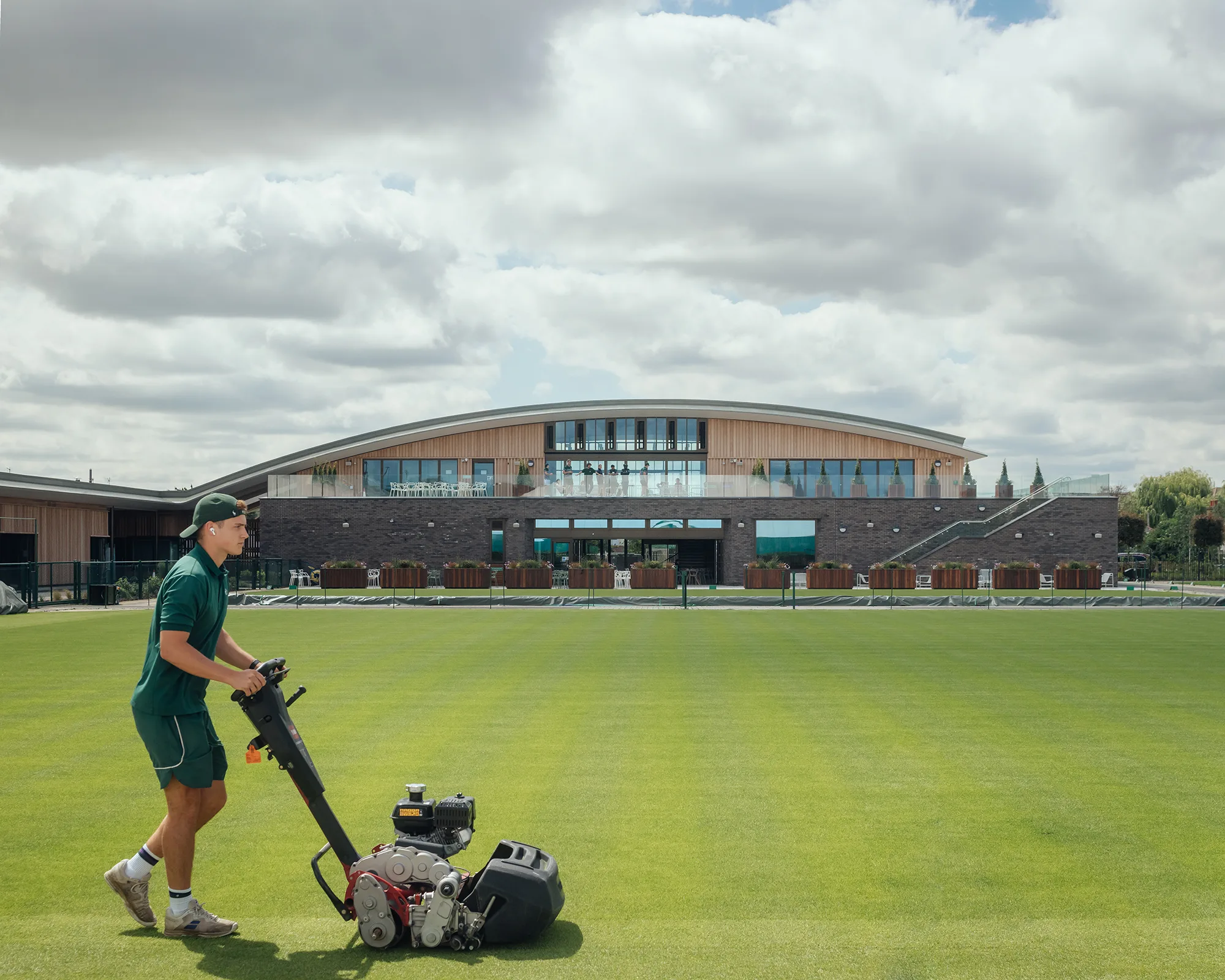 Tennis clubhouse in background with man in foreground cutting the grass