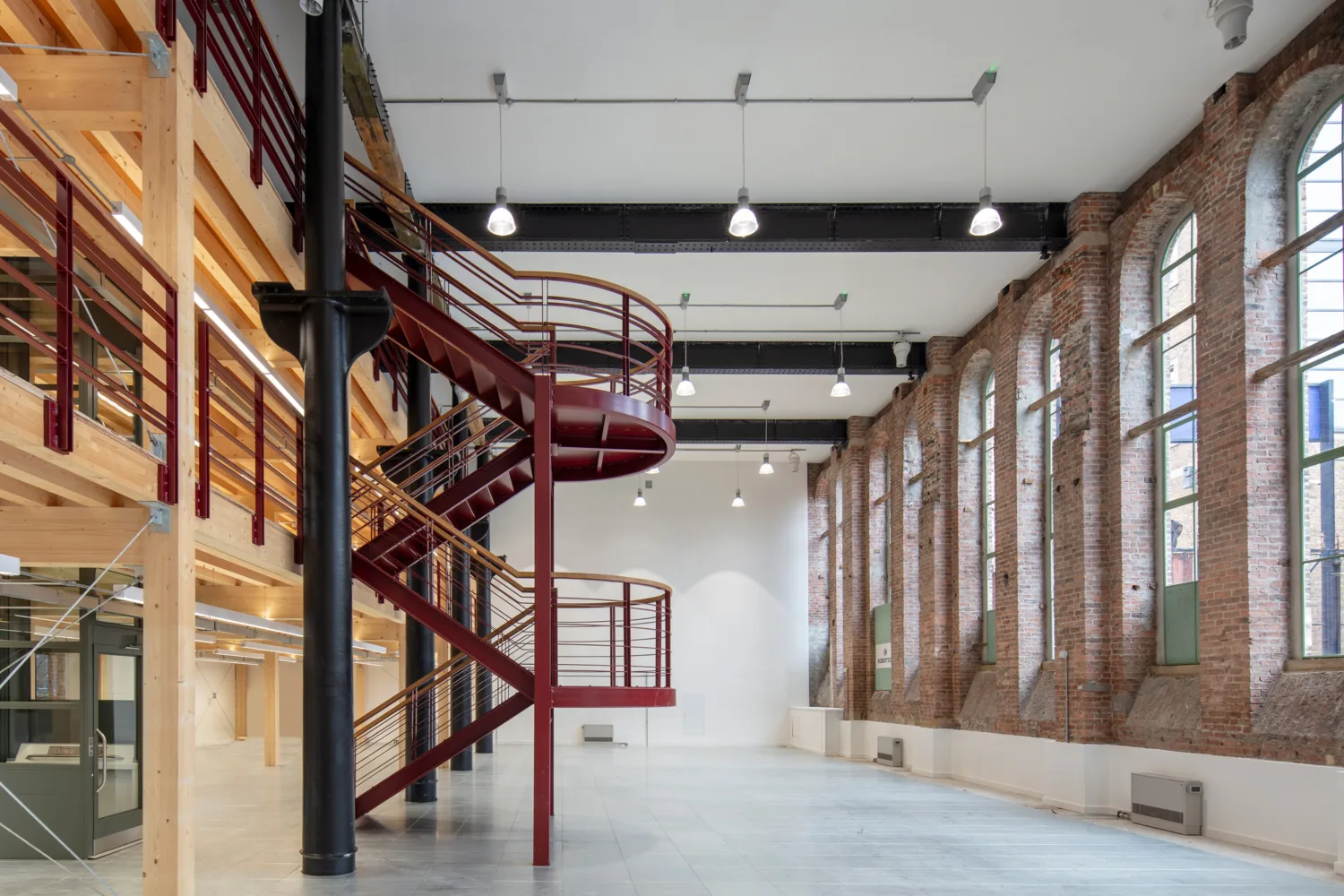 Indoor of building right hand side has red brick wall and large windows, there is a maroon staircase on the left leading to exposed levels.