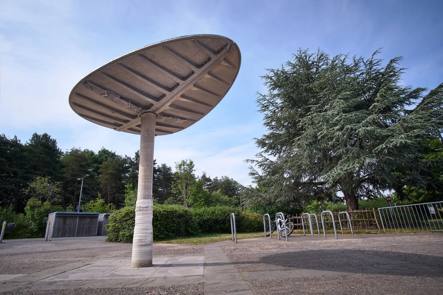 A large, concrete structure on a paved surface, with trees, a bike rack holding one bicycle, and a metal gate in the background under a clear blue sky.