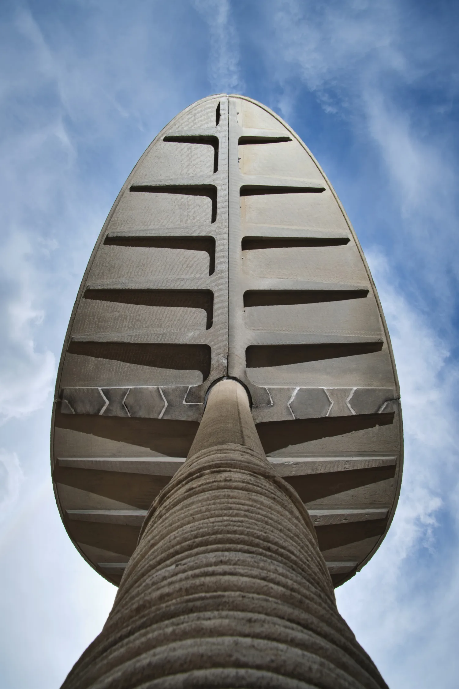 Low-angle view of the Canopy, a large concrete structure with a ribbed stem and segmented, oval-shaped top, set against a blue sky with scattered clouds.