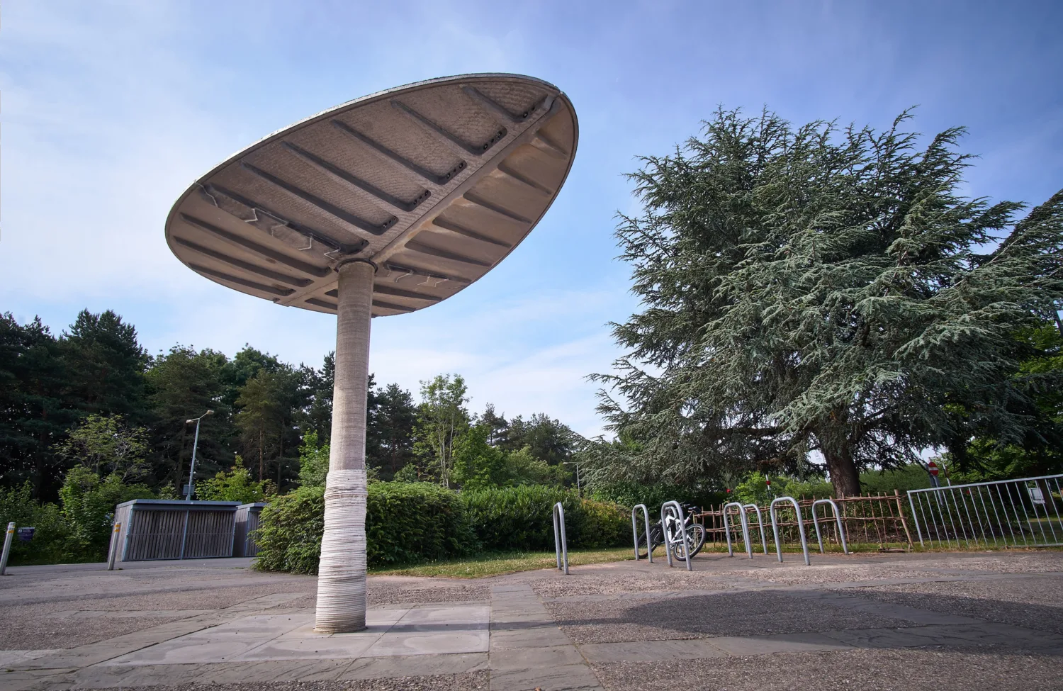 A large, concrete structure on a paved surface, with trees, a bike rack holding one bicycle, and a metal gate in the background under a clear blue sky.