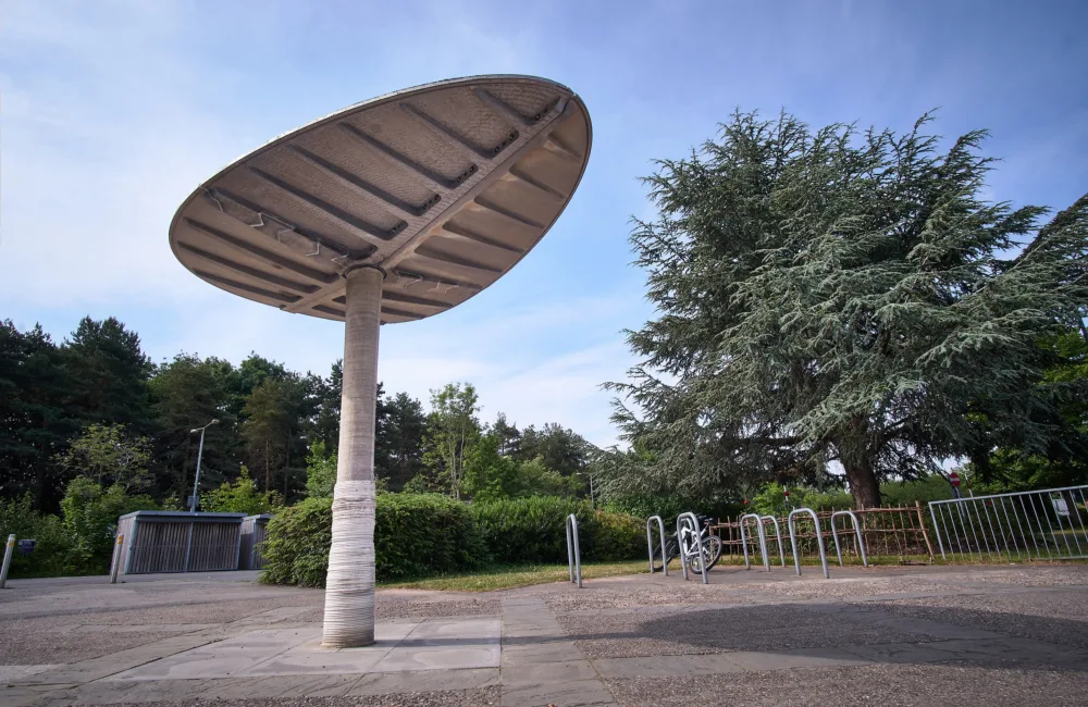 A large, concrete structure on a paved surface, with trees, a bike rack holding one bicycle, and a metal gate in the background under a clear blue sky.
