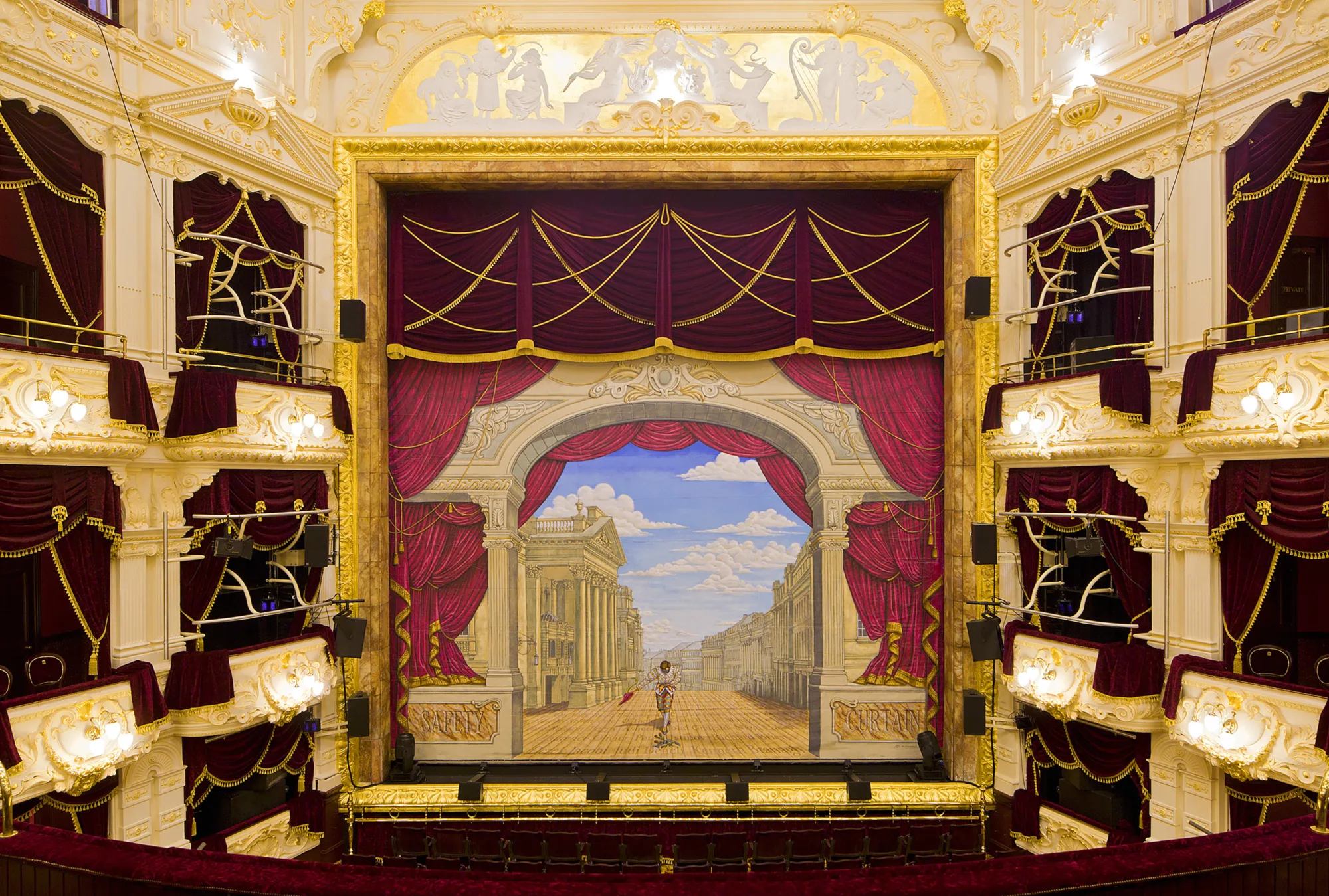 View of an ornate theatre stage framed by deep red curtains with gold trim, surrounded by multiple tiers of box seating with white and gold detailing. The backdrop shows a painted street scene under a blue sky.
