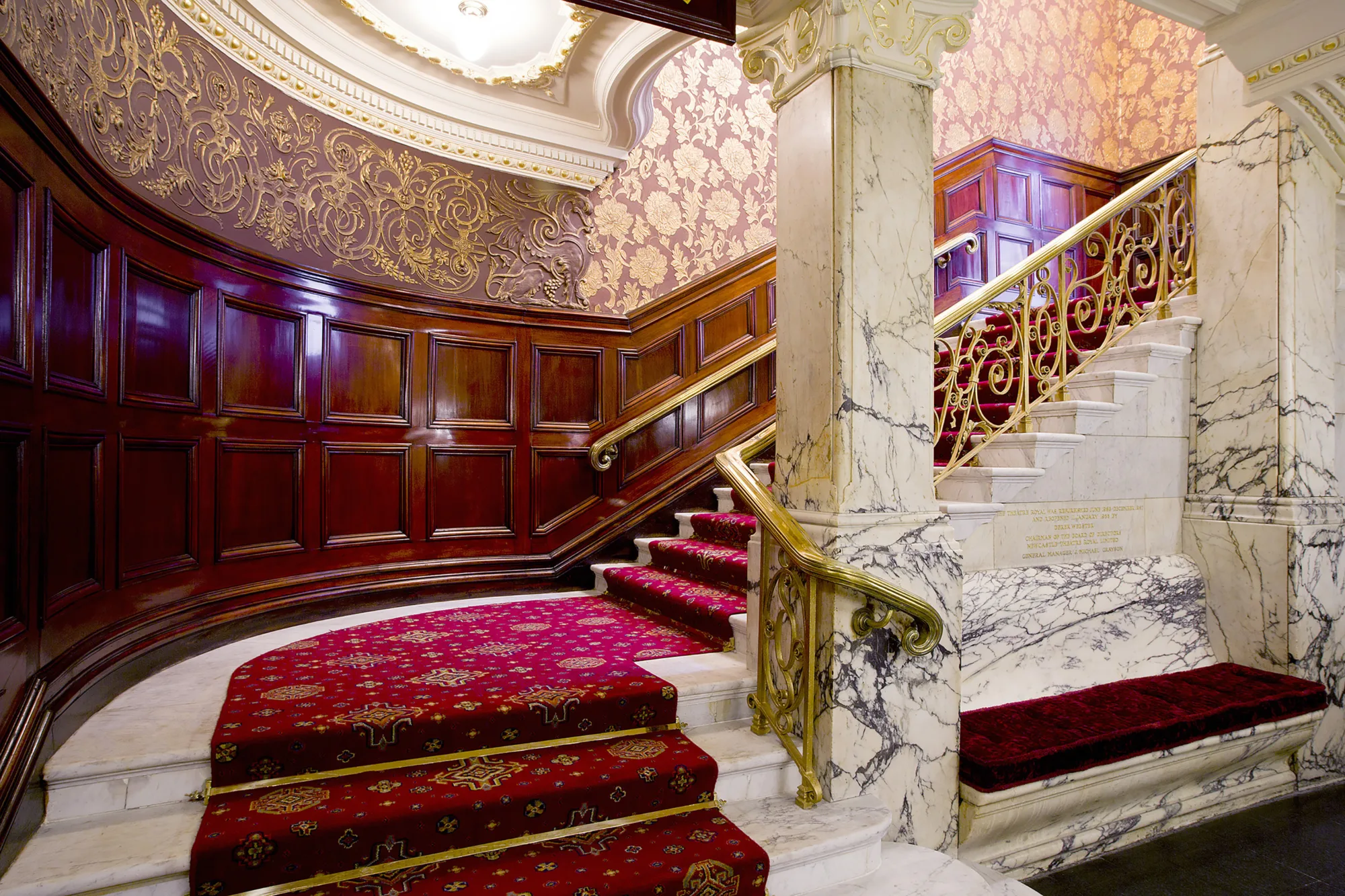 Curved staircase with red patterned carpet and white marble steps, featuring ornate gold handrails and decorative panels. Dark wood wall paneling and red damask wallpaper with gold detailing surround the area.