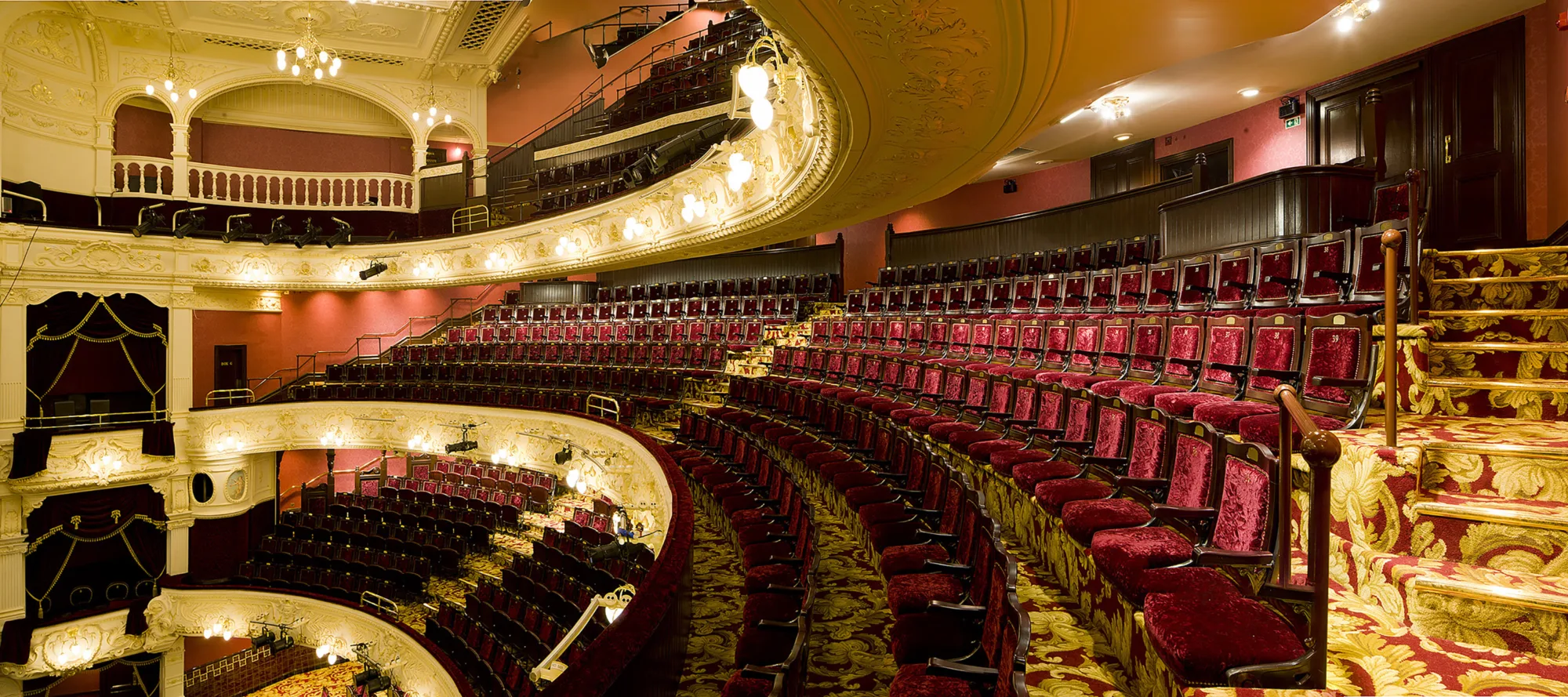 Curved rows of red velvet seats in a multi-tiered theatre balcony with ornate white and gold detailing. Patterned carpet covers steps leading to upper seating areas under warm lighting.