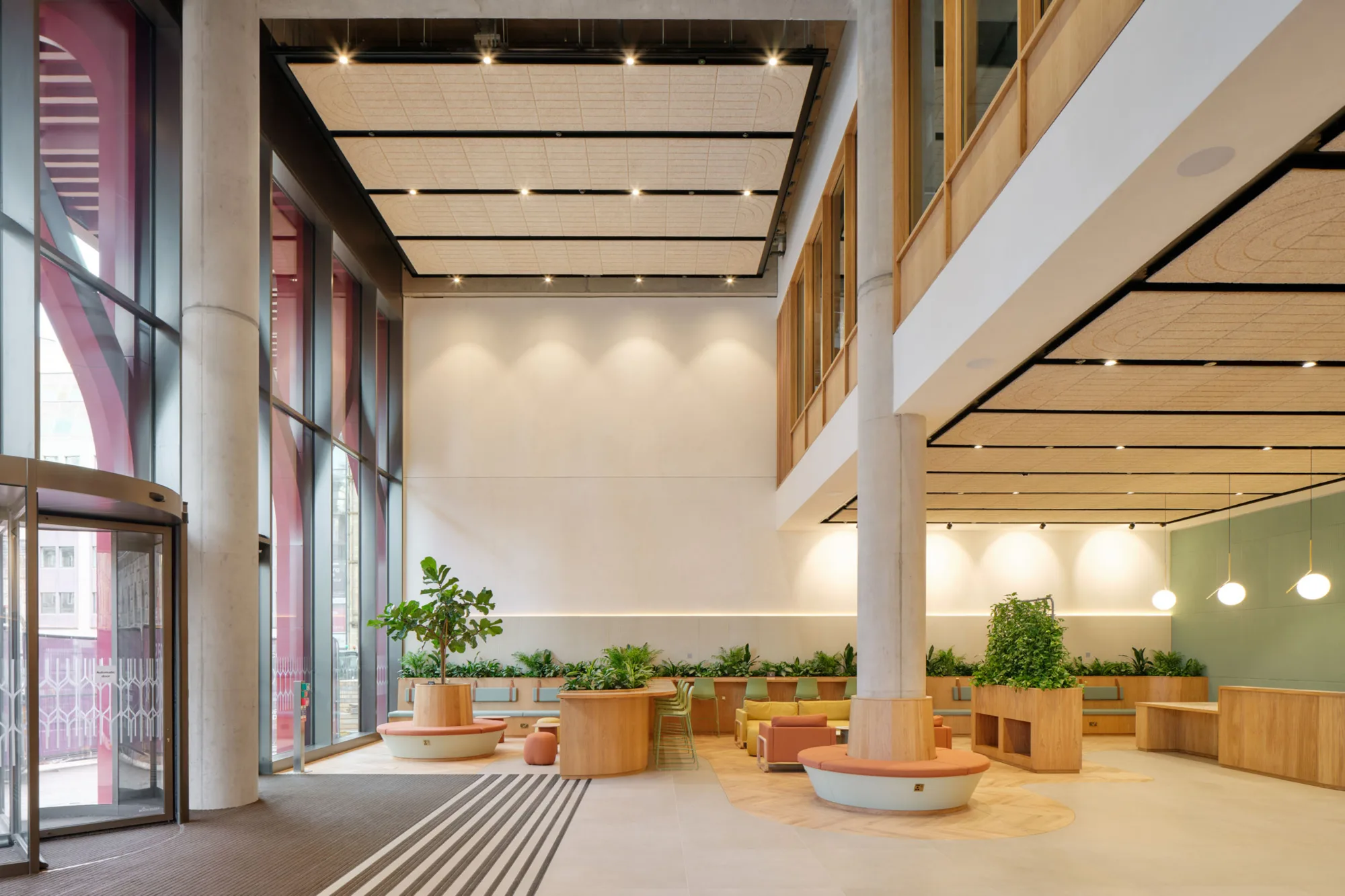 Spacious modern lobby with tall glass windows, light-colored flooring, and wooden seating areas arranged around planters with green plants. Ceiling panels with integrated lighting and a revolving glass door are visible on the left, with additional seating and pendant lights in the background.