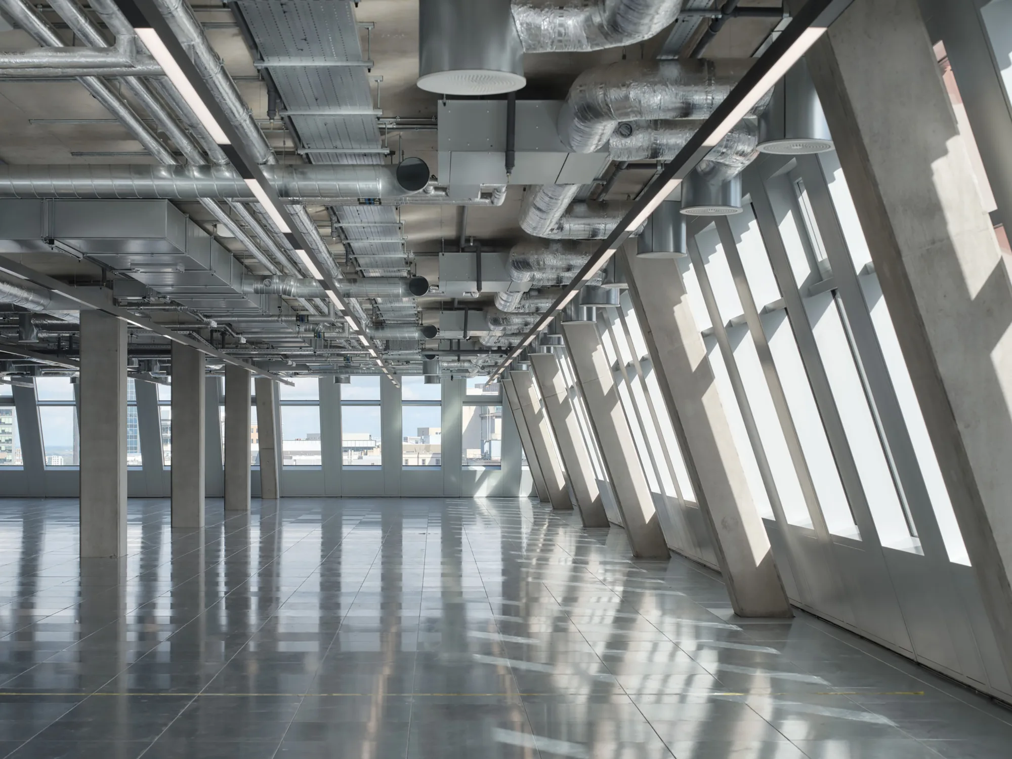 Interior view of an empty commercial space with large angled windows on the right, polished reflective flooring, and exposed ceiling featuring metal ducts, pipes, and lighting fixtures. Concrete columns run along the left side, and city buildings are visible through the windows.