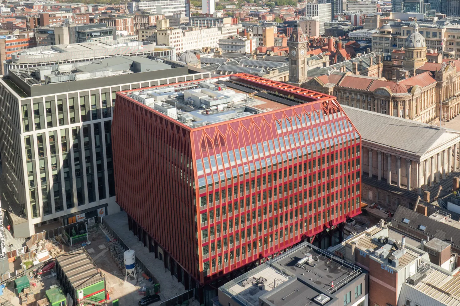 Aerial view of a city block featuring a modern red building with a slanted roof and vertical fins, surrounded by older historic structures and other office buildings. Rooftop equipment is visible, and streets with construction areas are in the foreground.