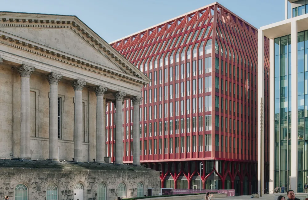 modern red building with vertical fins and a slanted roof on the right. A glass-fronted building is partially visible on the far right, with pedestrians walking in the foreground.