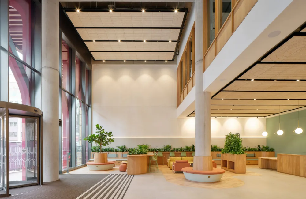 Spacious modern lobby with tall glass windows, light-colored flooring, and wooden seating areas arranged around planters with green plants. Ceiling panels with integrated lighting and a revolving glass door are visible on the left, with additional seating and pendant lights in the background.