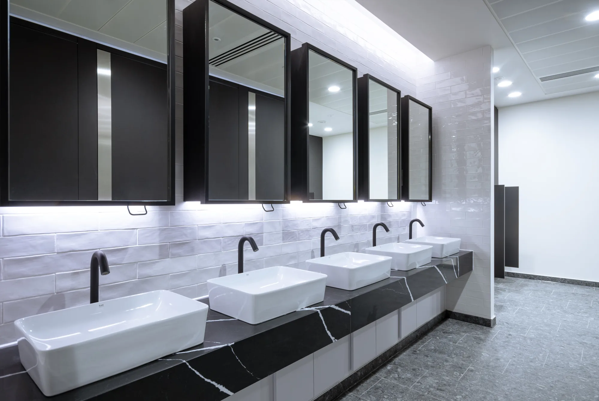 Internal view of white bathrom sinks above grey and white marbled surface, above is a row of mirrors and a grey floor