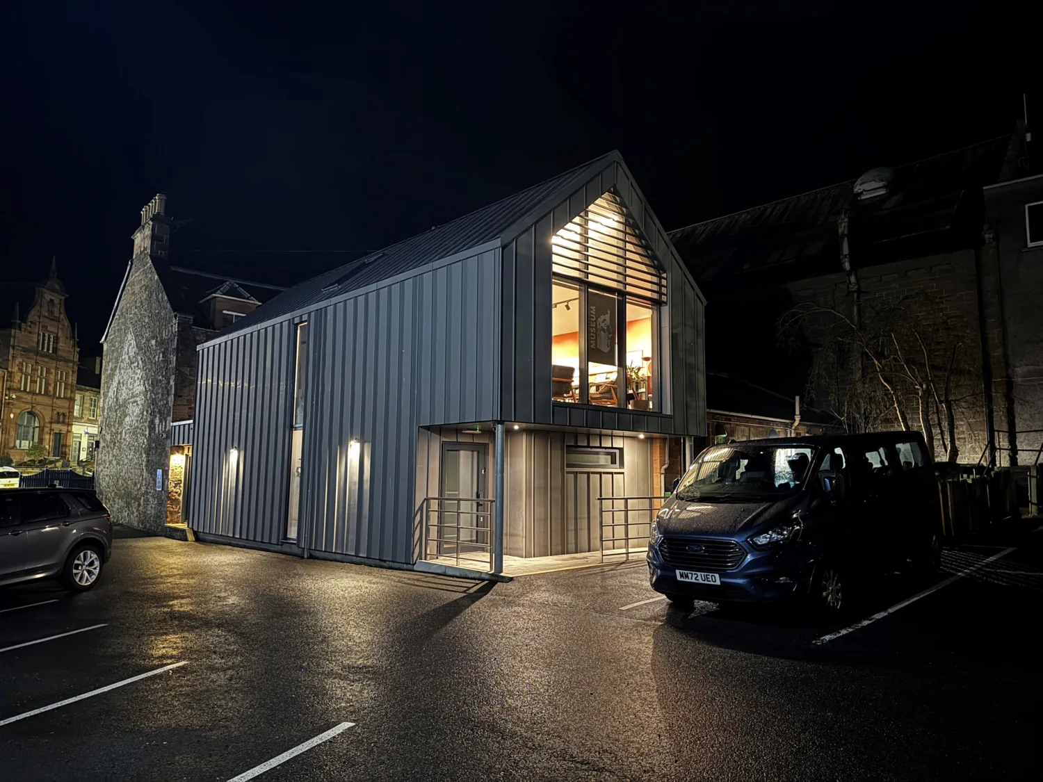 Nighttime view of a modern two‑storey metal‑clad building with a large illuminated upper window, set beside older stone buildings and parked vehicles in a wet parking lot.