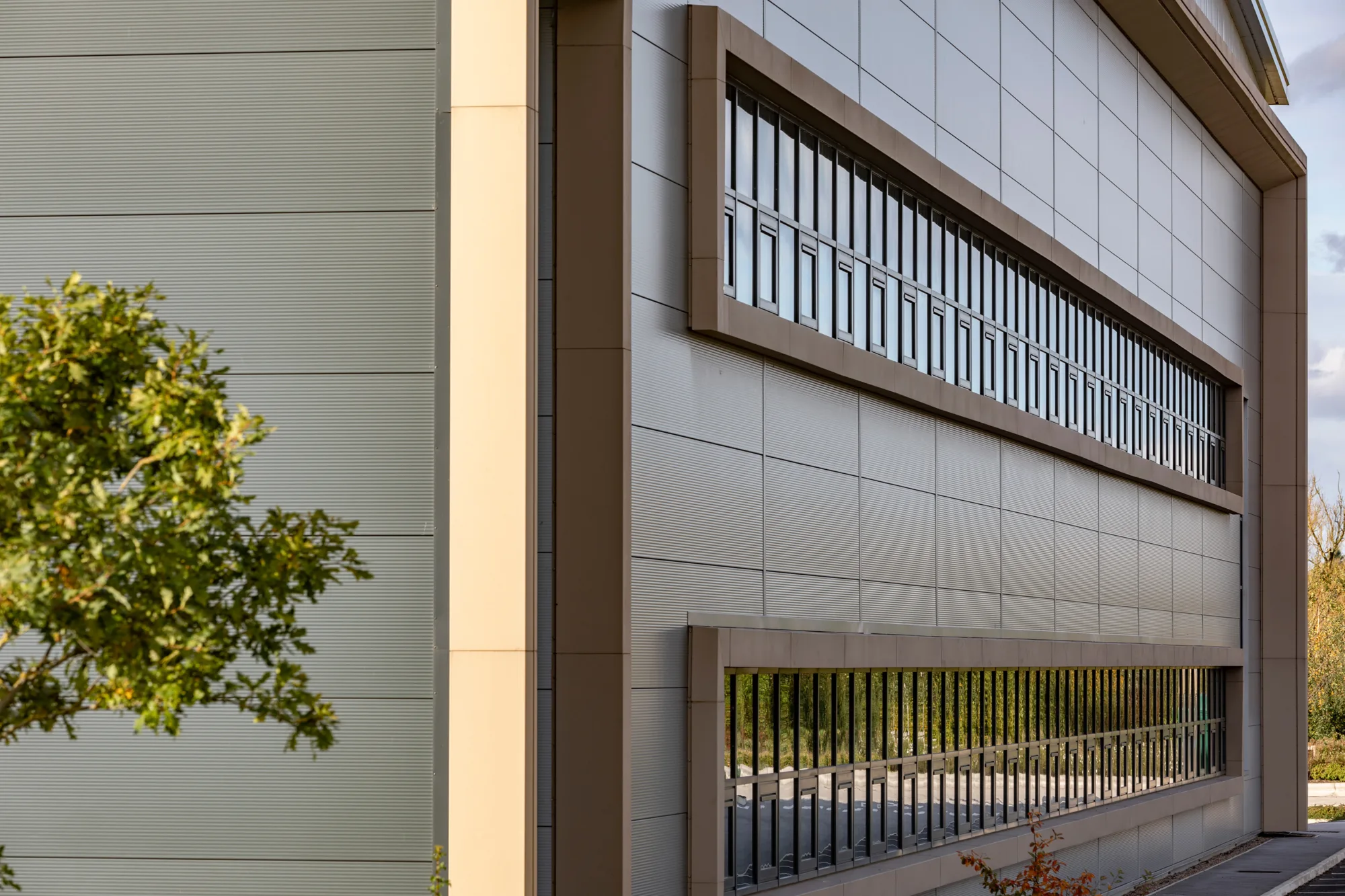 Modern industrial building with beige and grey exterior, two horizontal rows of windows and a tree with green leaves on the left side.