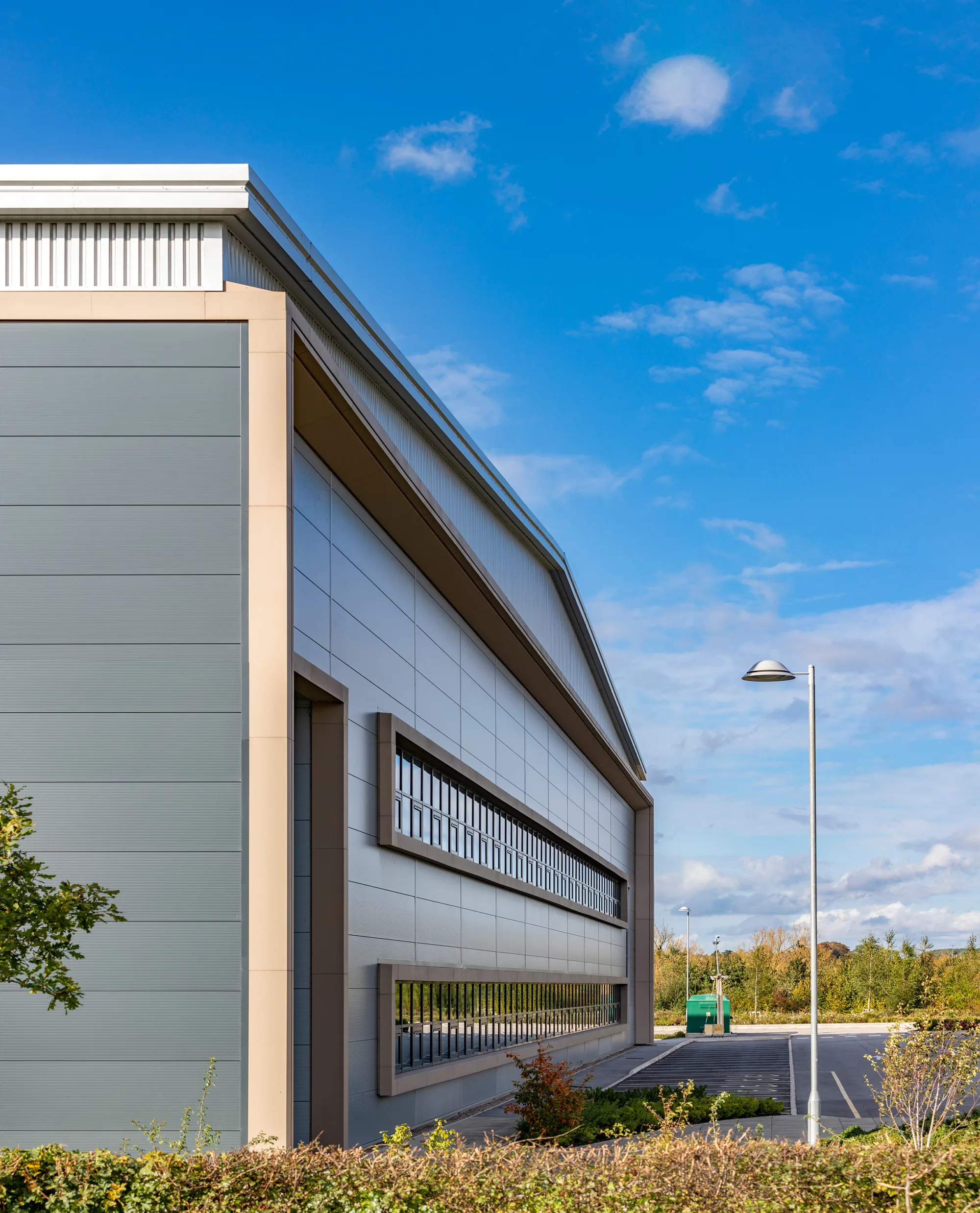 Modern industrial building with grey and beige exterior, large horizontal windows, surrounded by greenery and a parking lot with a streetlamp. Sky is clear with scattered clouds.