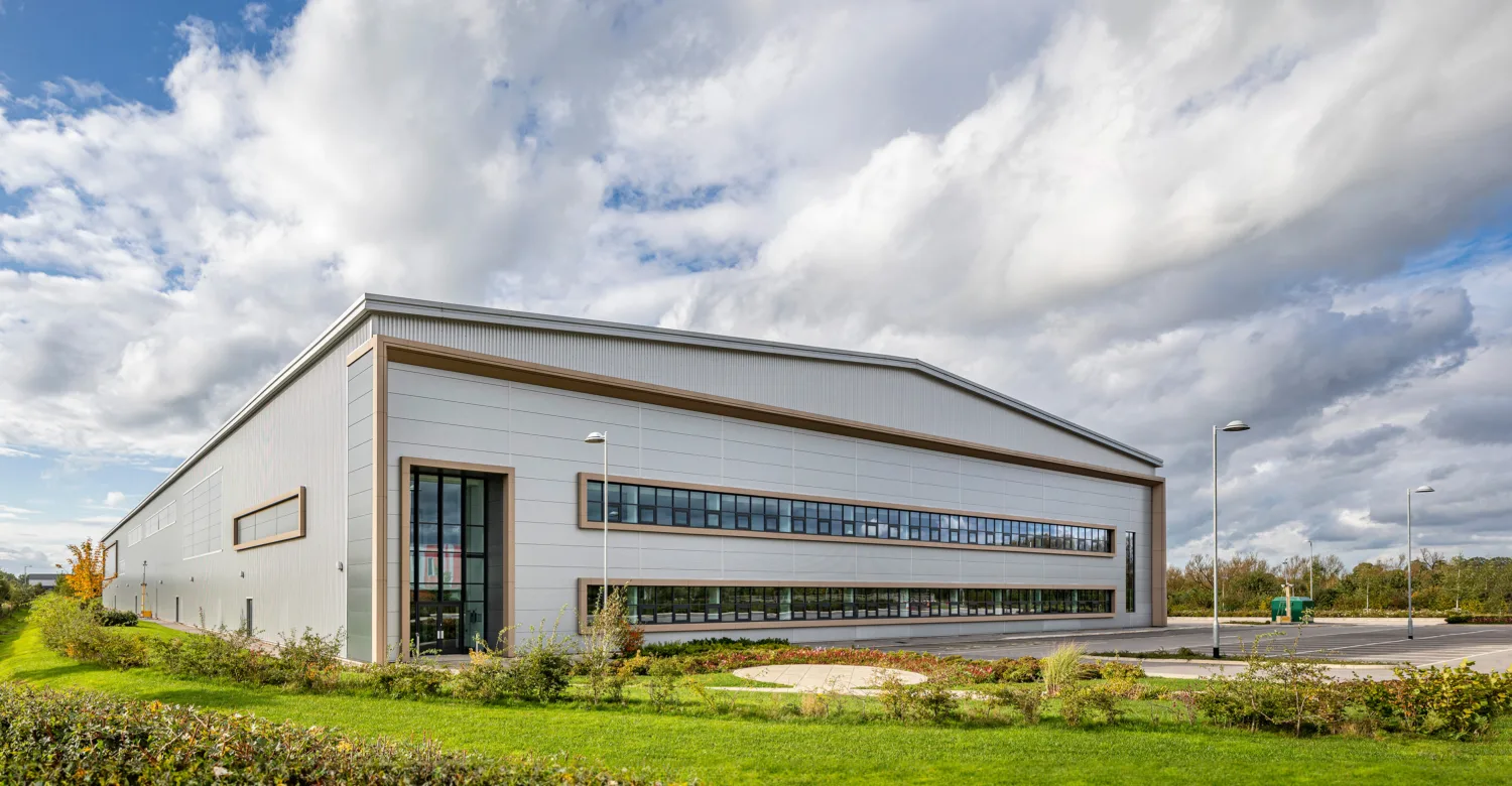 Large modern industrial building with light grey exterior and horizontal windows, surrounded by lawn and bushes. Cloudy sky above, streetlights and empty parking lot on the right.