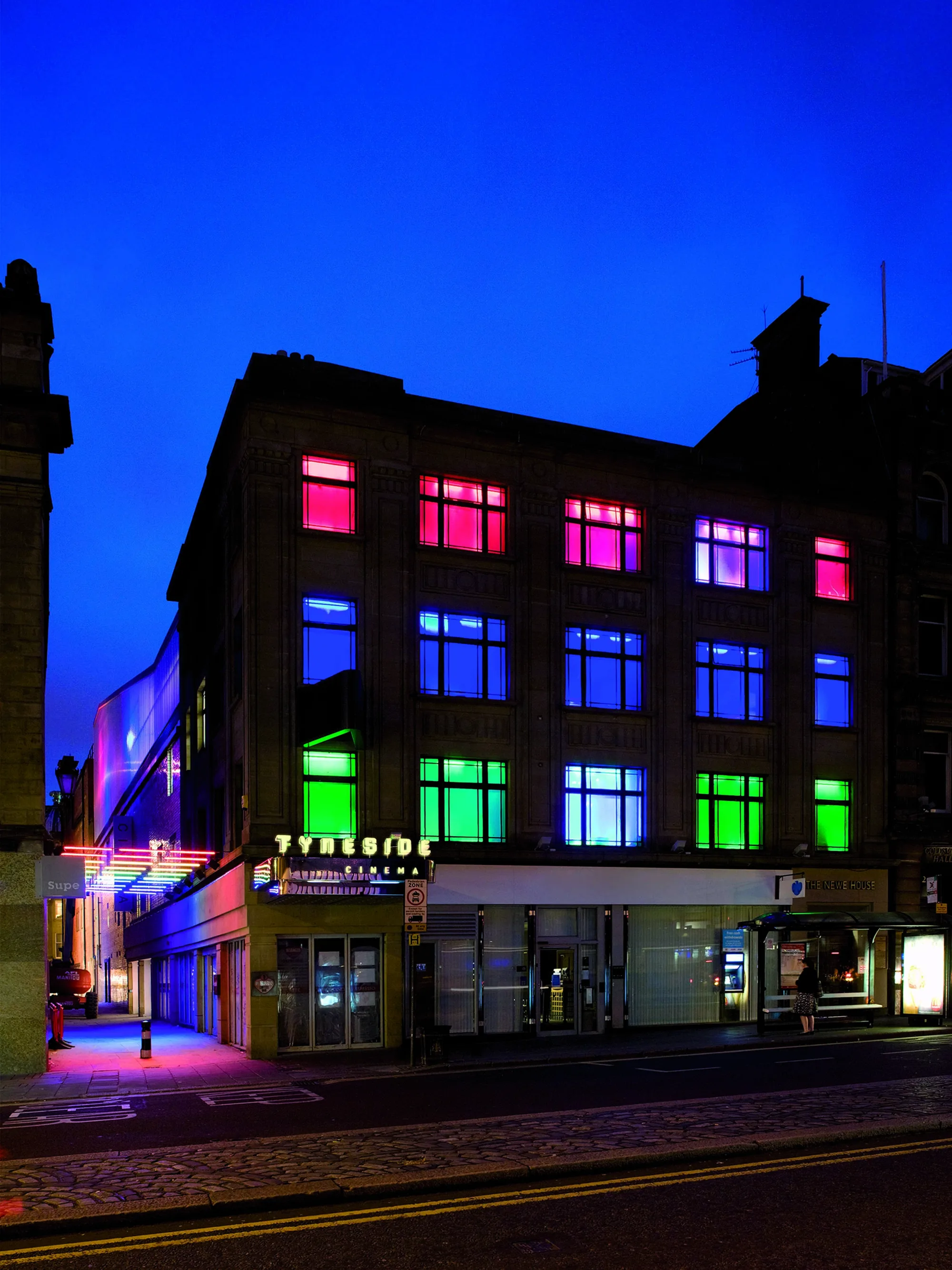 External view of facade of Cinema, its twilight and the windows are light in red, blue, green and purple, there is an alleyway to the left lt by neon