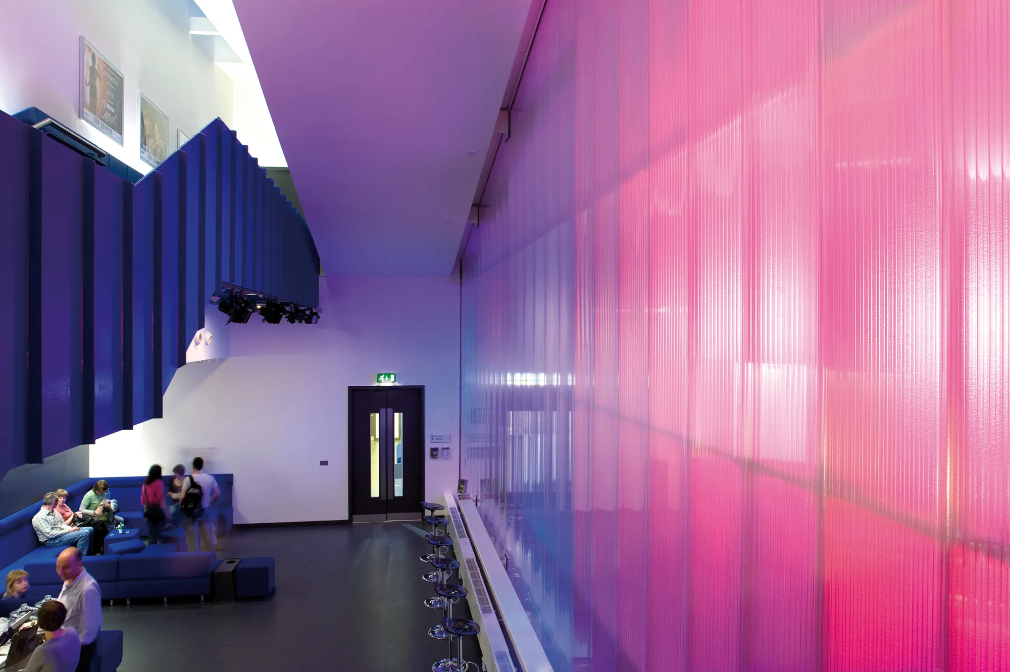 shot of bar area, there is a pinky purply wall on the right hand side, there is an elevaated blue clad staircase on the left and people seated on blue sofas underneath.