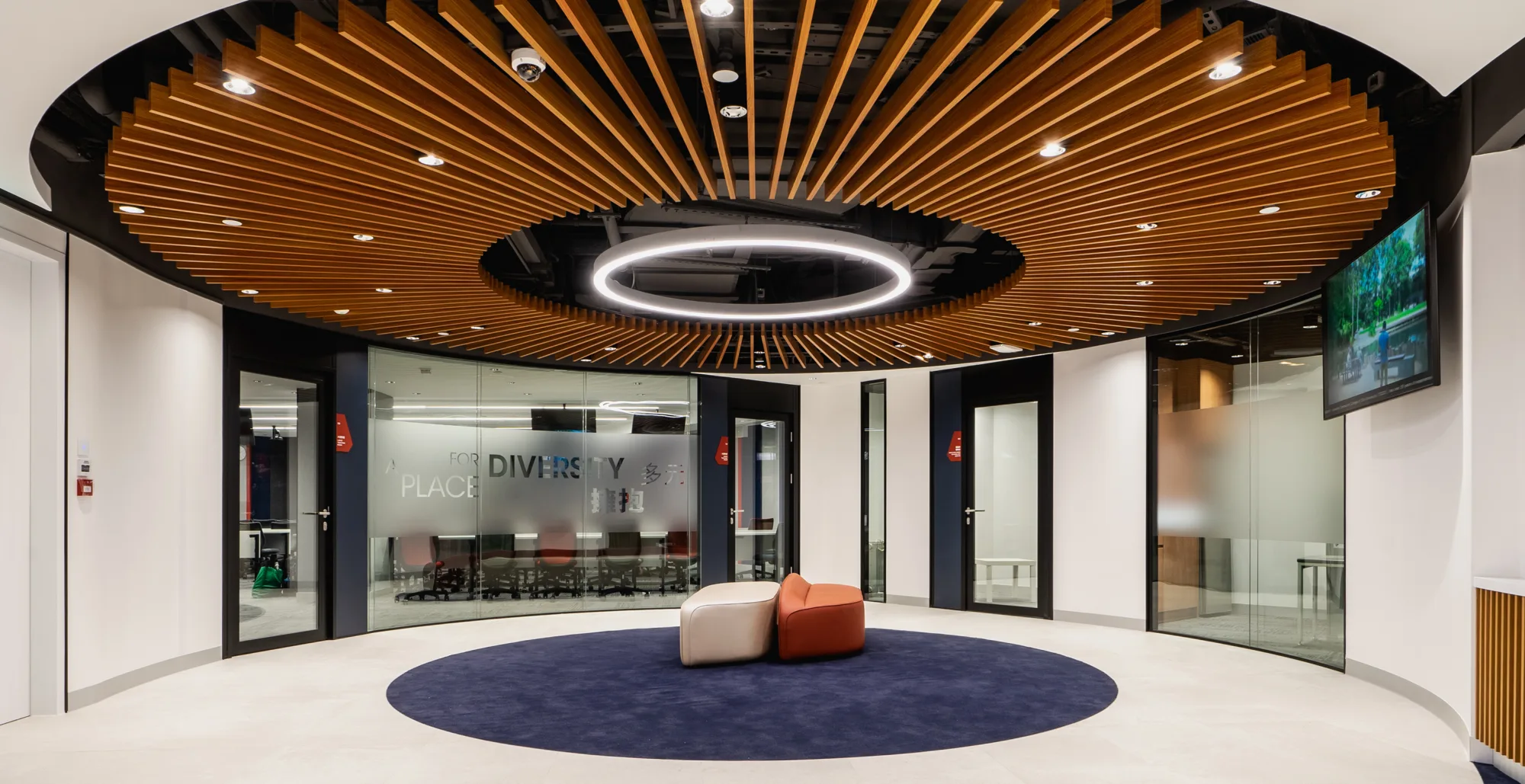 Rotunda with adjacent sofas in the centre underneath circular LED light; features TV screen on wall and glass partitions