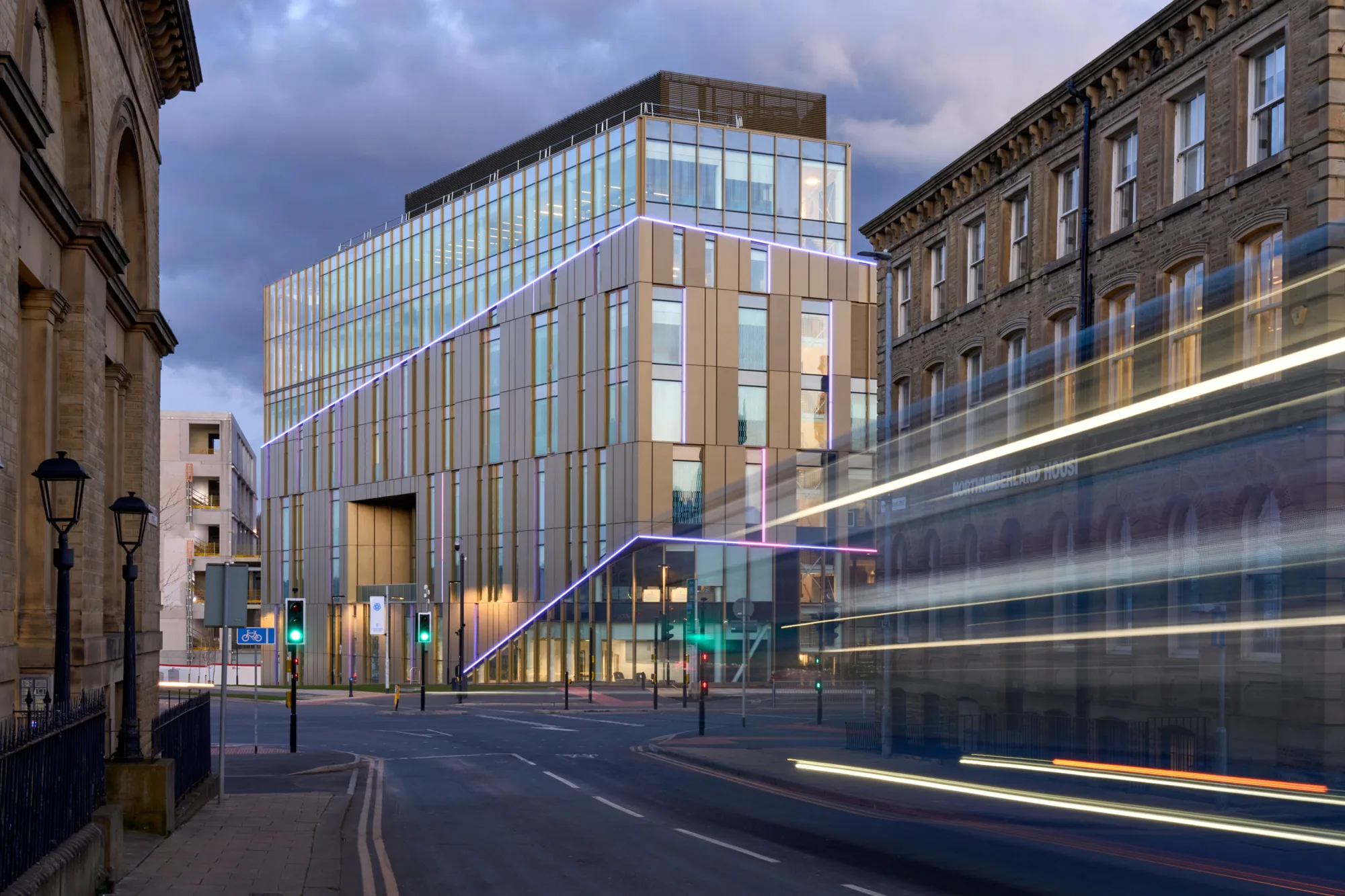 A multi-story building with glass windows and vertical beige panels, outlined by purple lights, stands at a street intersection; a blurred bus moves on the right side of the image.